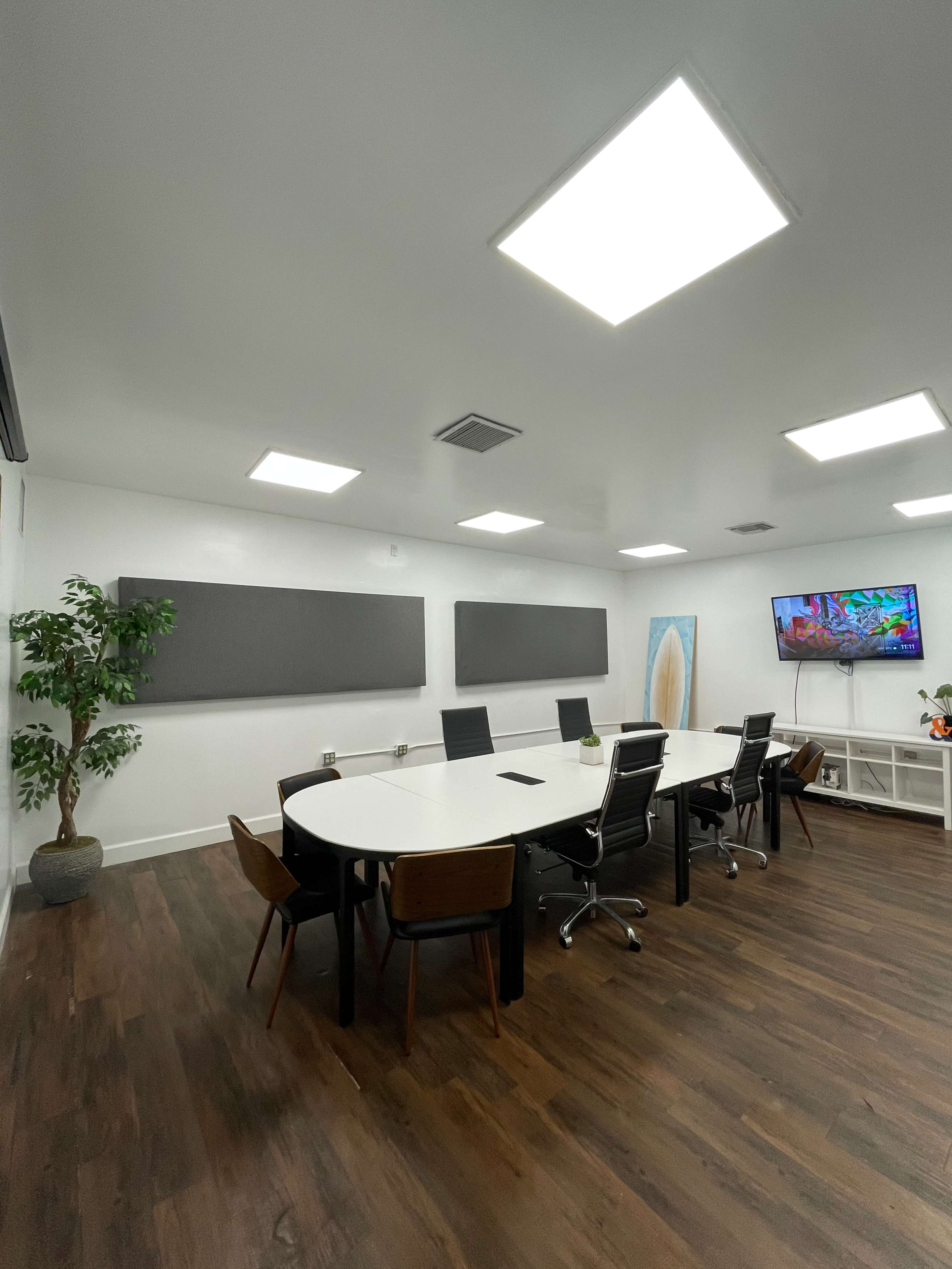 A modern conference room with a large oval table surrounded by black chairs, a TV on the wall, and two sound-absorbing panels flanking the space.