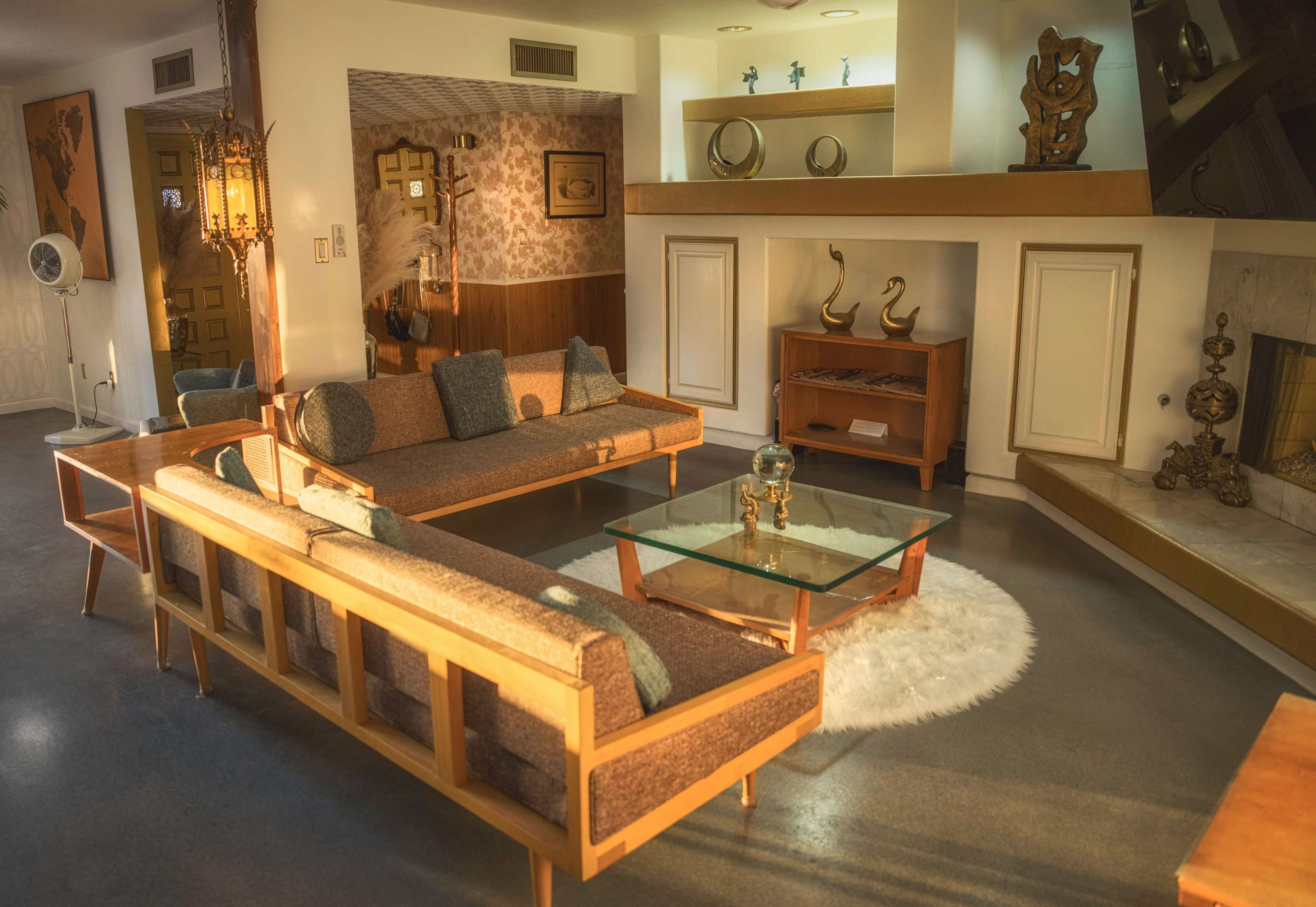 The image shows a mid-century modern living room featuring a pair of wooden framed sofas, a glass coffee table on a round rug, and a fireplace with decorative items.