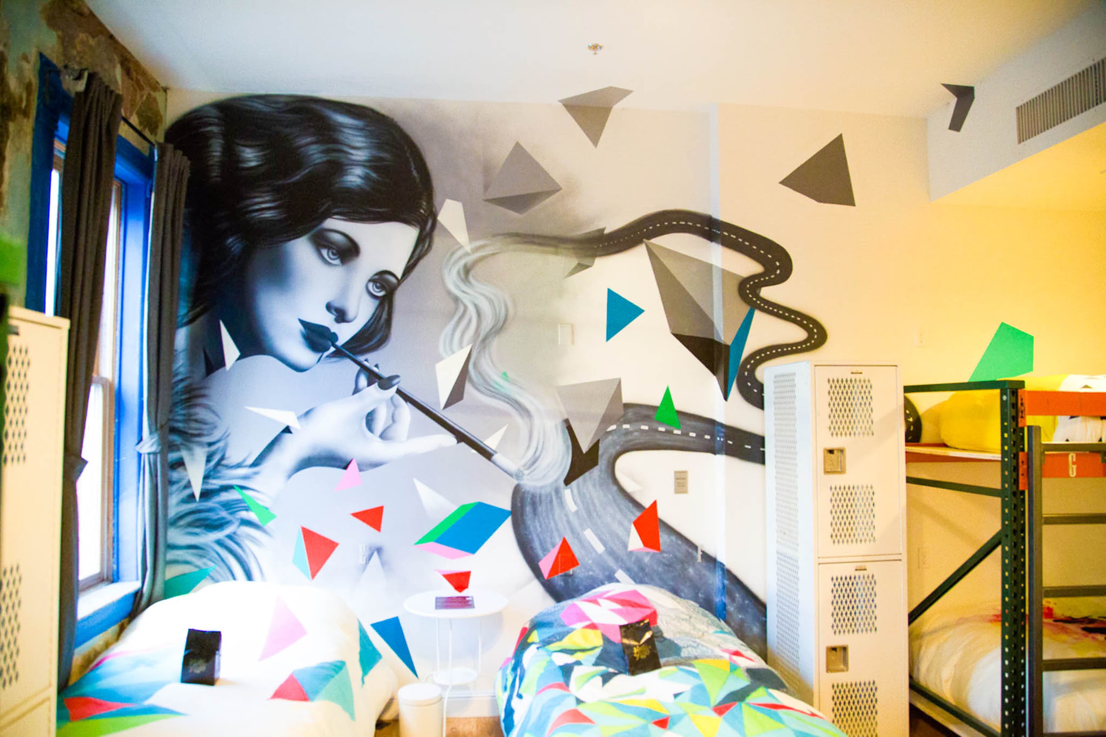 The image shows a room with two beds and a large mural of a woman playing a flute, surrounded by colorful geometric shapes on the walls.