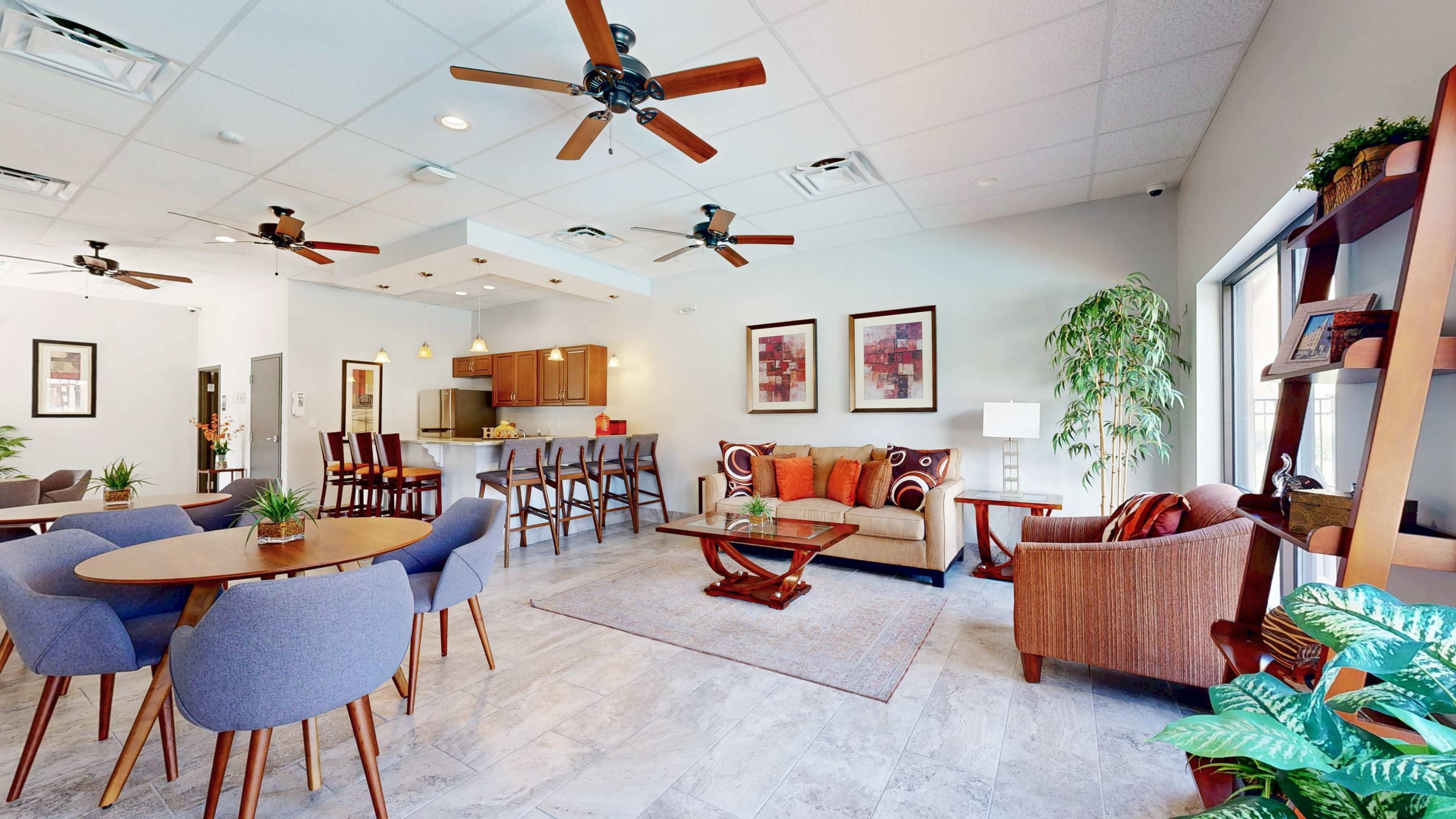 The image shows a modern common living area featuring a seating arrangement with a sofa, chairs, and dining tables, alongside a kitchenette area.
