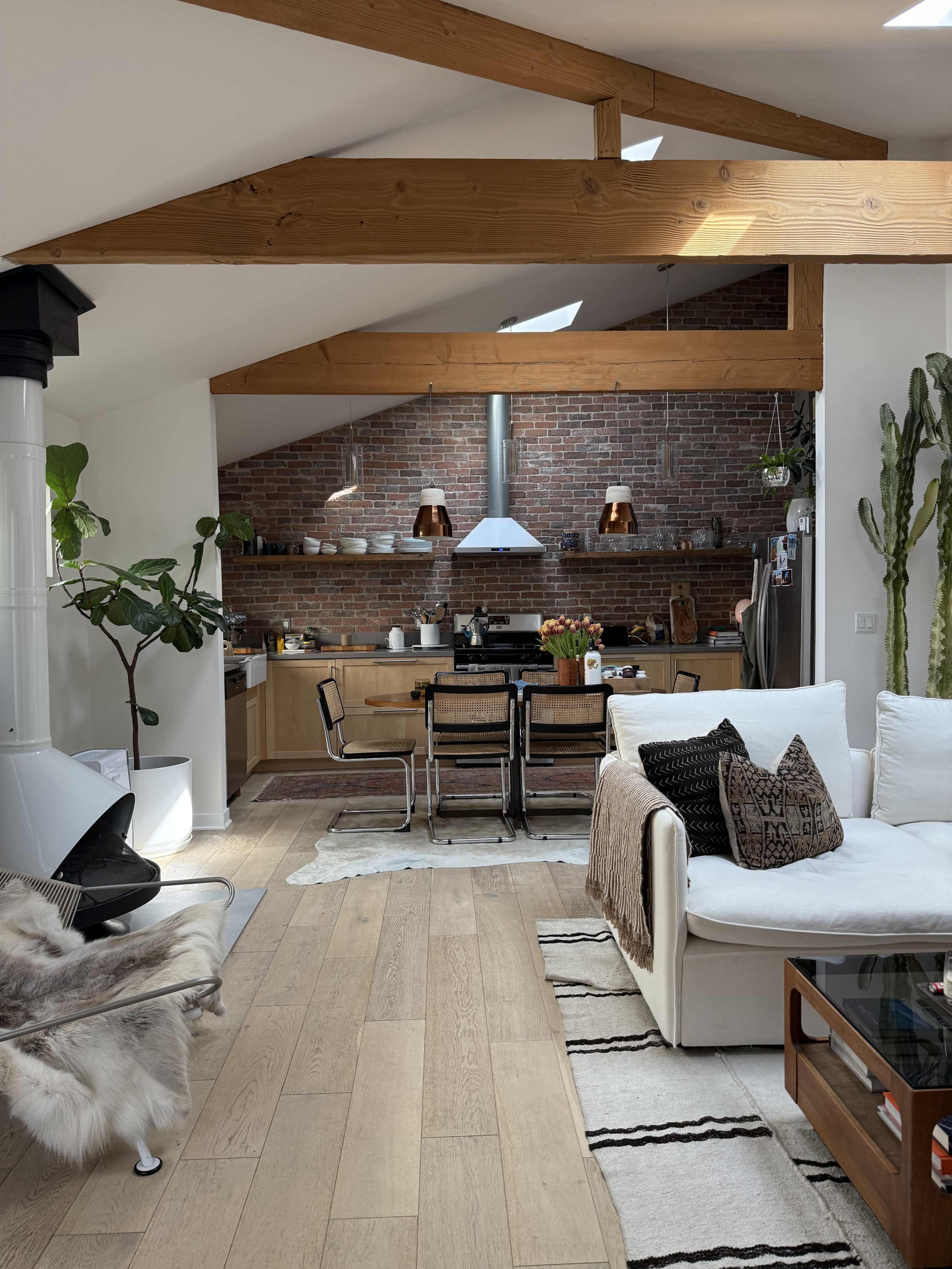 The image shows a spacious, modern kitchen and living area with wooden beams, brick walls, and a mix of contemporary furniture.