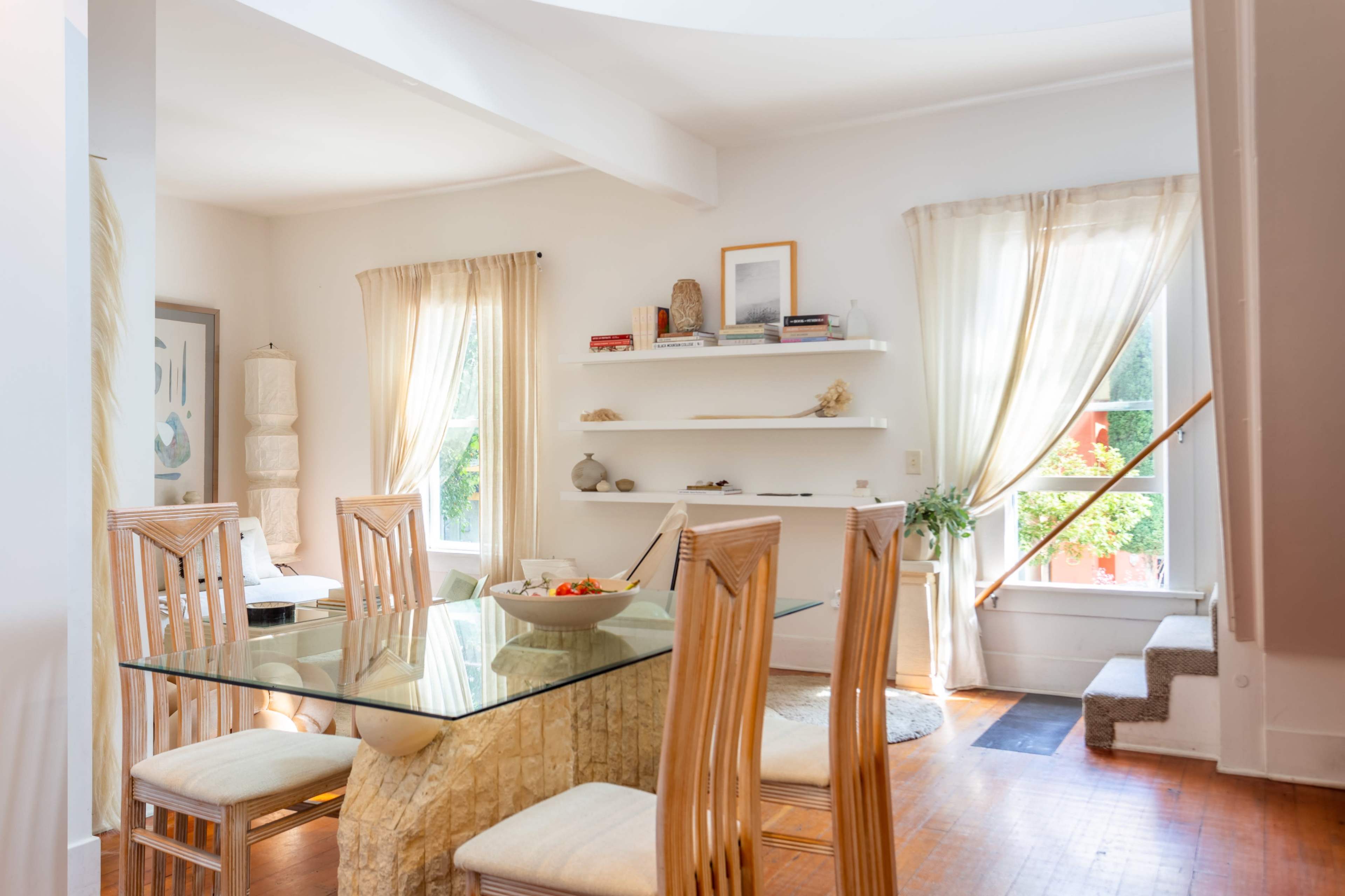 A dining area features a glass table surrounded by wooden chairs, with shelves on the wall displaying decorative items and large windows allowing natural light.