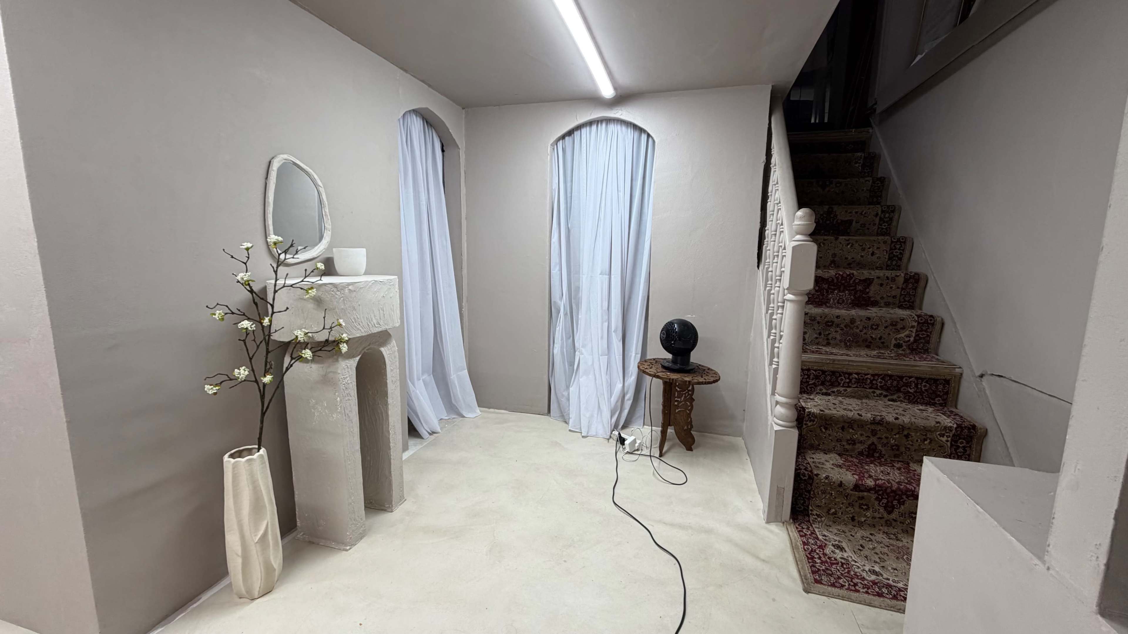 The image shows a light-colored hallway with a staircase on the right, a decorative mantelpiece, a plant in a vase, and a black lamp on a small wooden table.