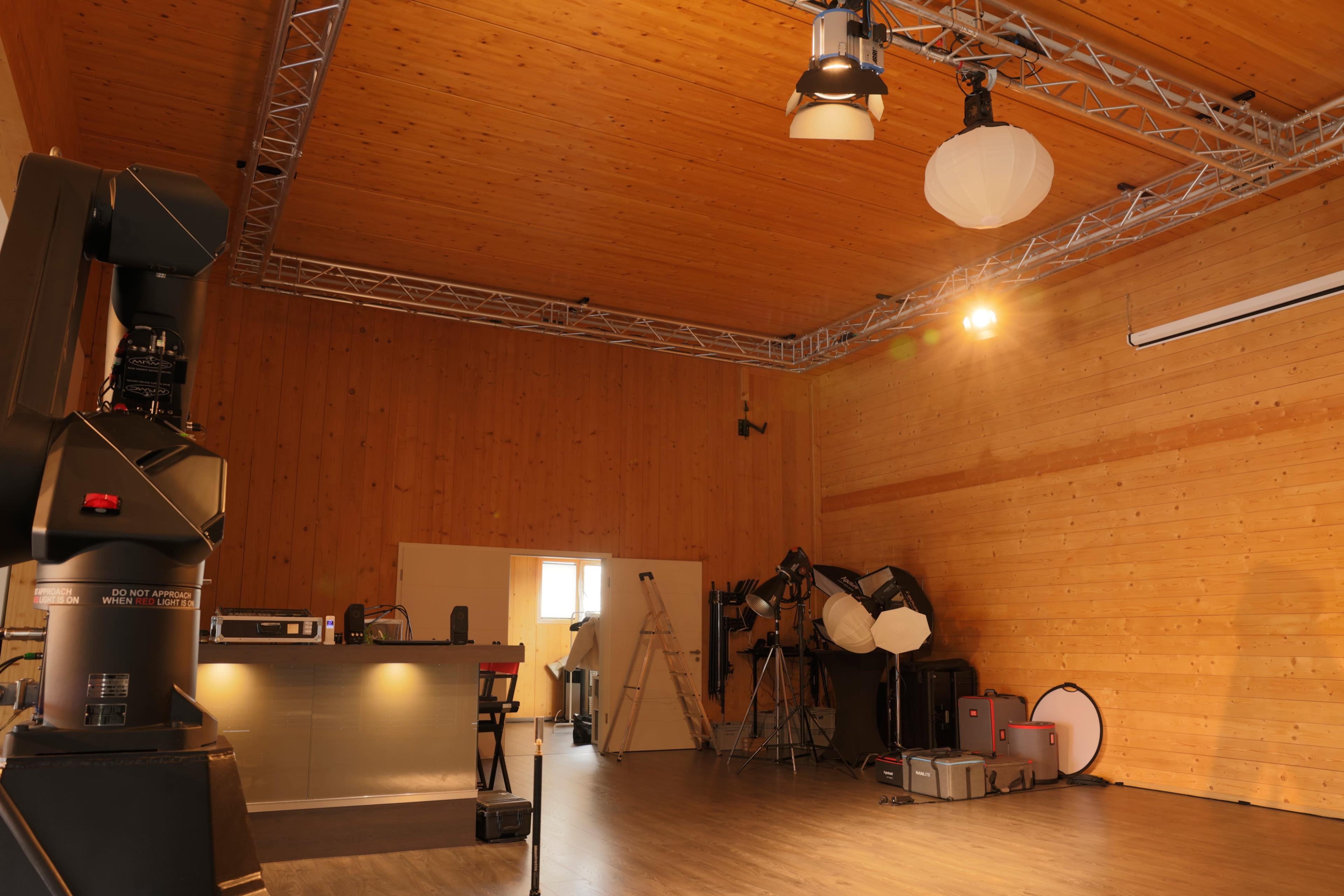 The image depicts a spacious interior studio with wooden walls, various lighting equipment, and a kitchenette area in the background.