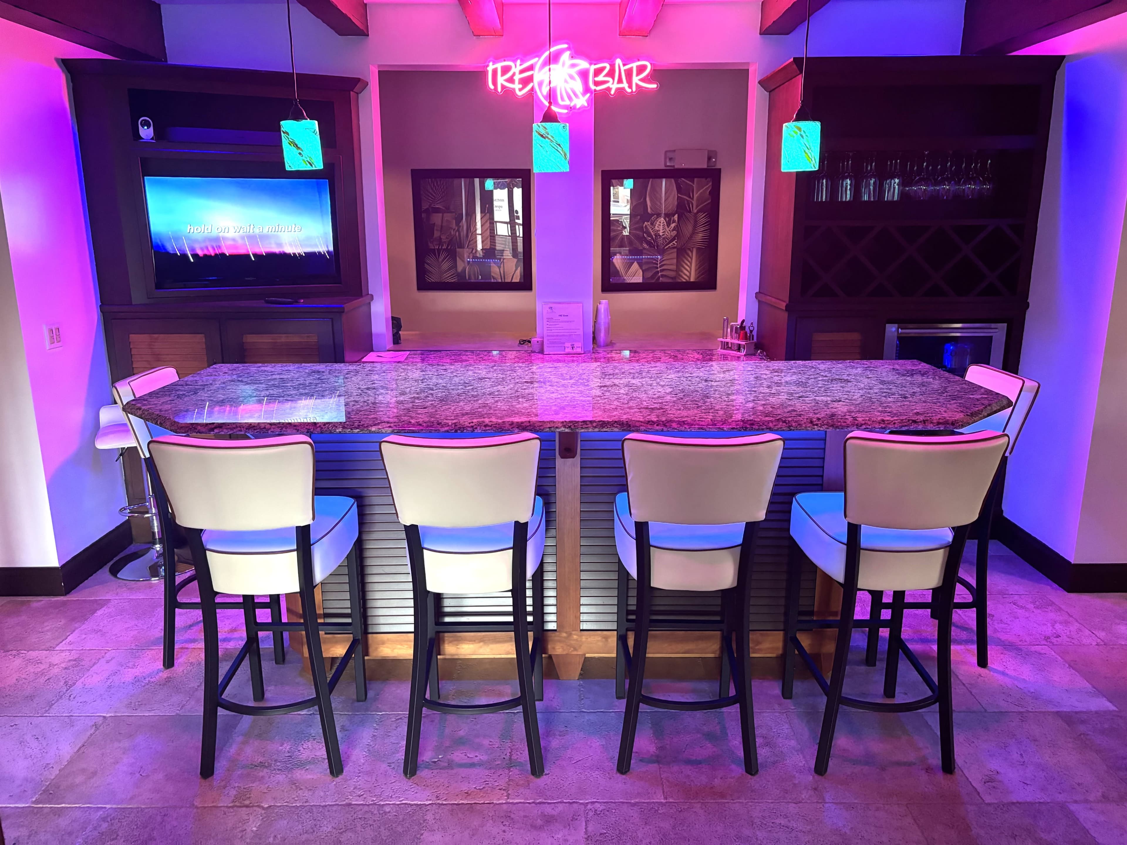 A modern bar area with a granite countertop, five white bar stools, and illuminated decorations including a neon "Tiki Bar" sign.