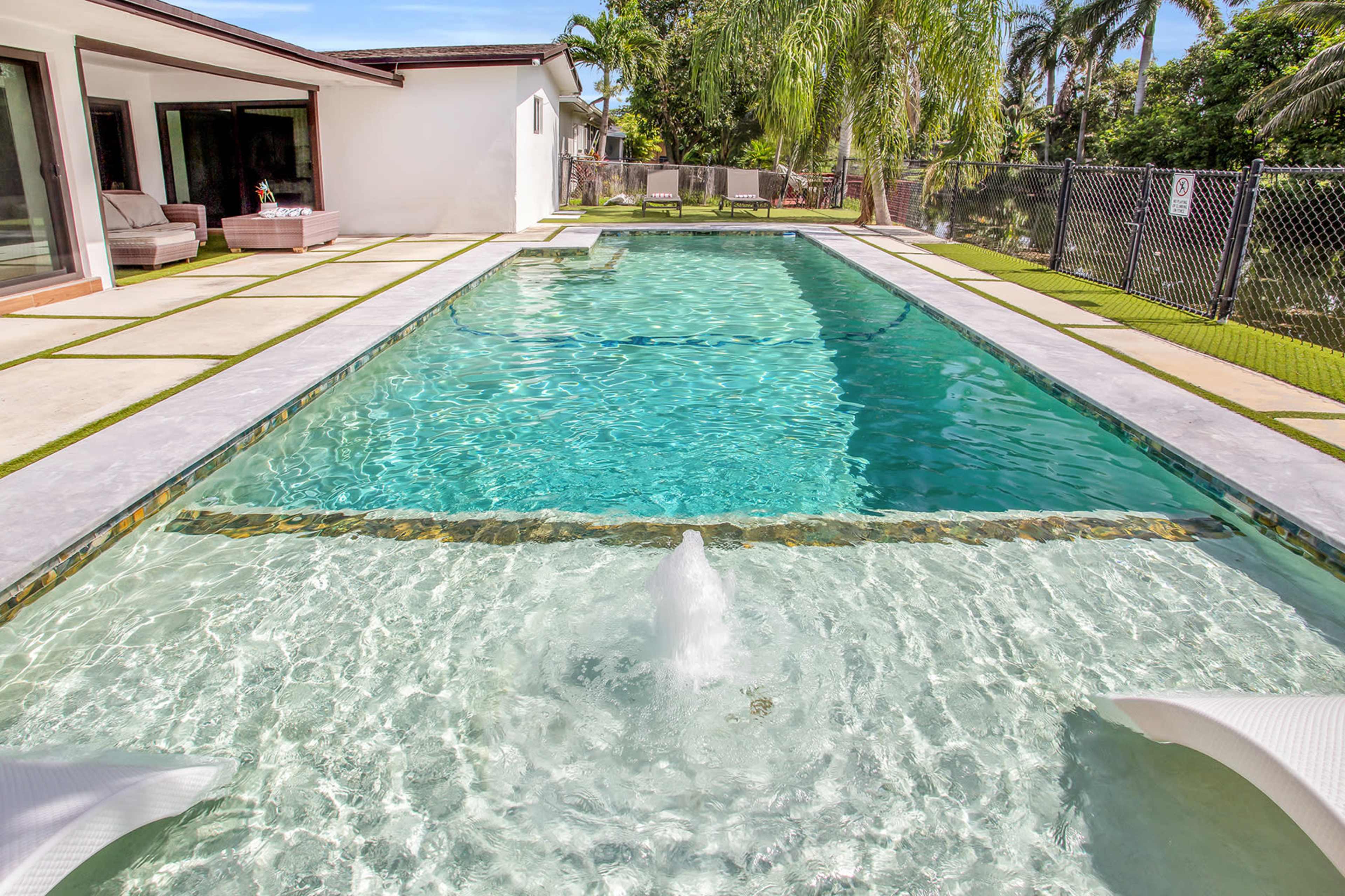 The image shows a swimming pool surrounded by a paved deck and landscaped area, with lounge chairs and palm trees in the background.