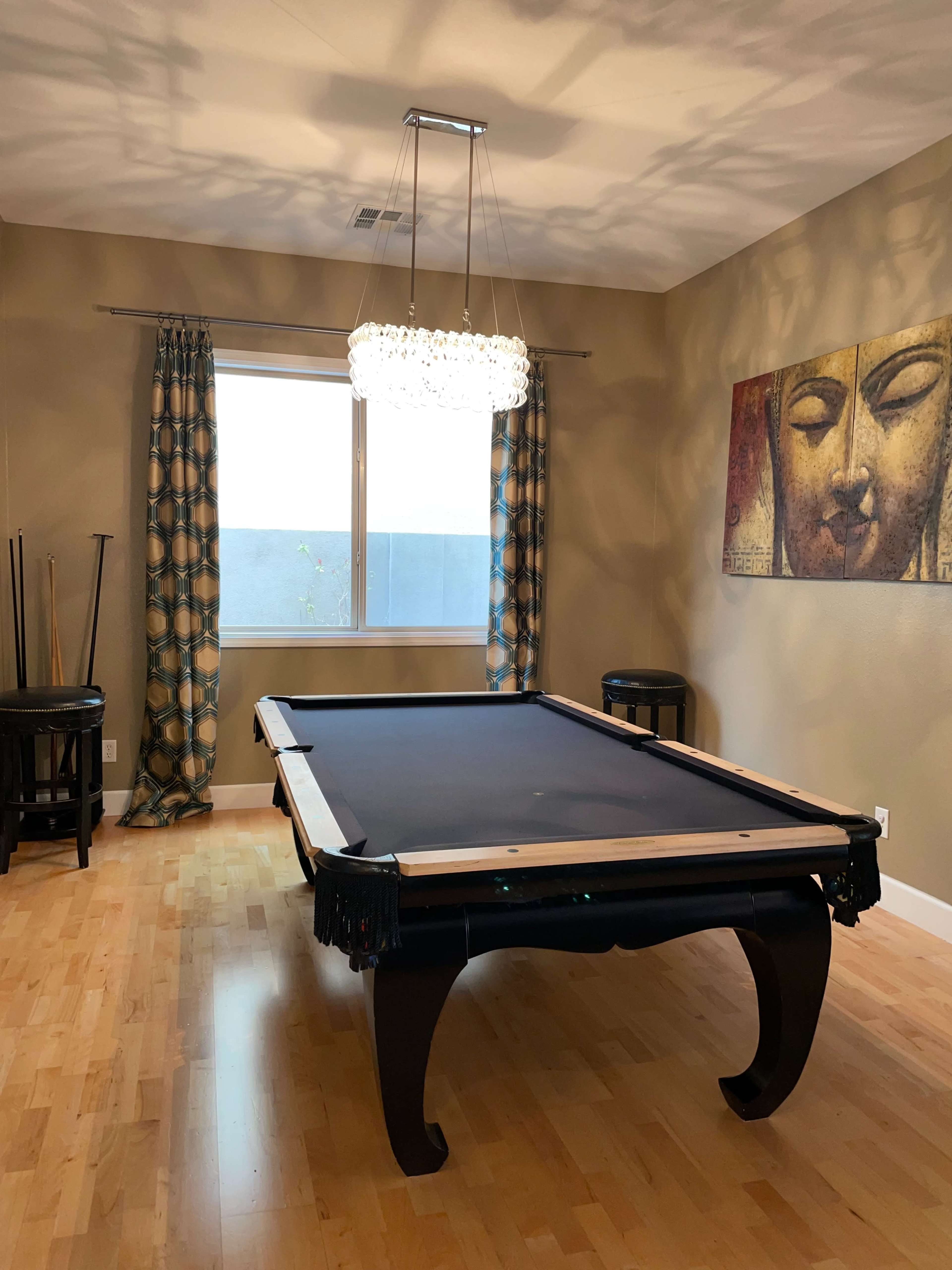 A well-lit room featuring a pool table, a modern light fixture, and a large window with decorative curtains.