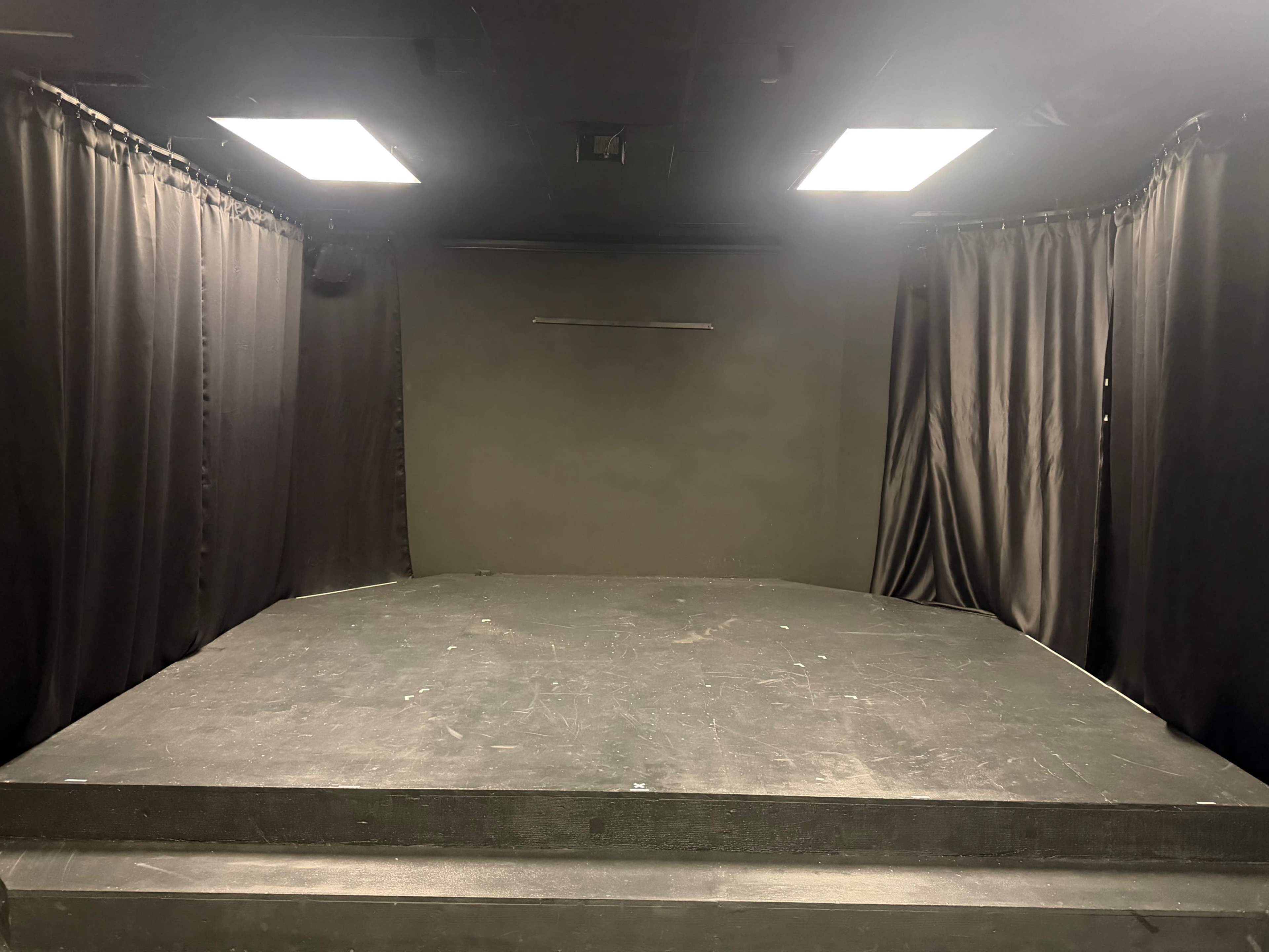 The image shows a small stage set with a dark backdrop, two upright curtains on either side, and overhead lighting.
