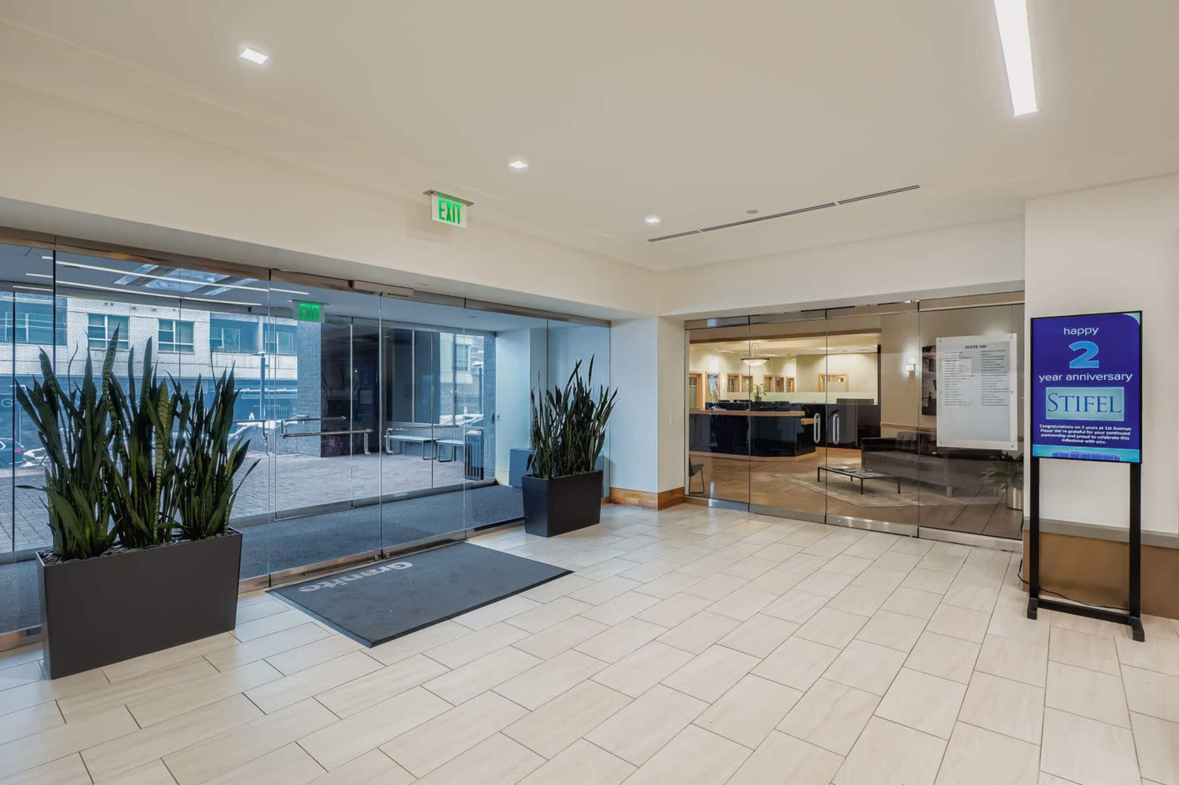 The image shows a modern office lobby with large glass doors, potted plants on either side, and a sign promoting a two-year anniversary celebration.