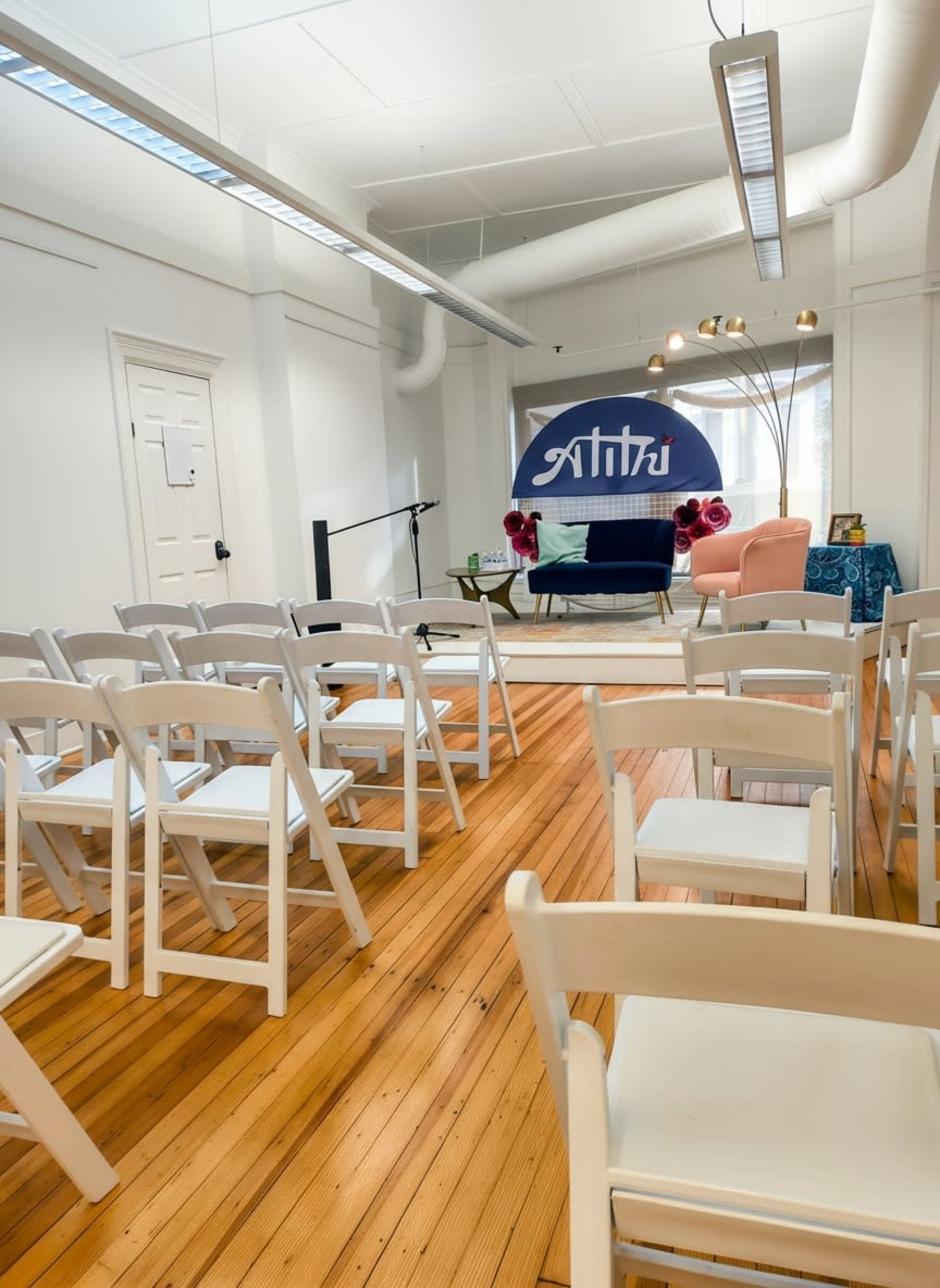 The image shows a room arranged with white chairs facing a small stage featuring a couch, a table, and decorative elements, with a large backdrop displaying the word "Anthū."