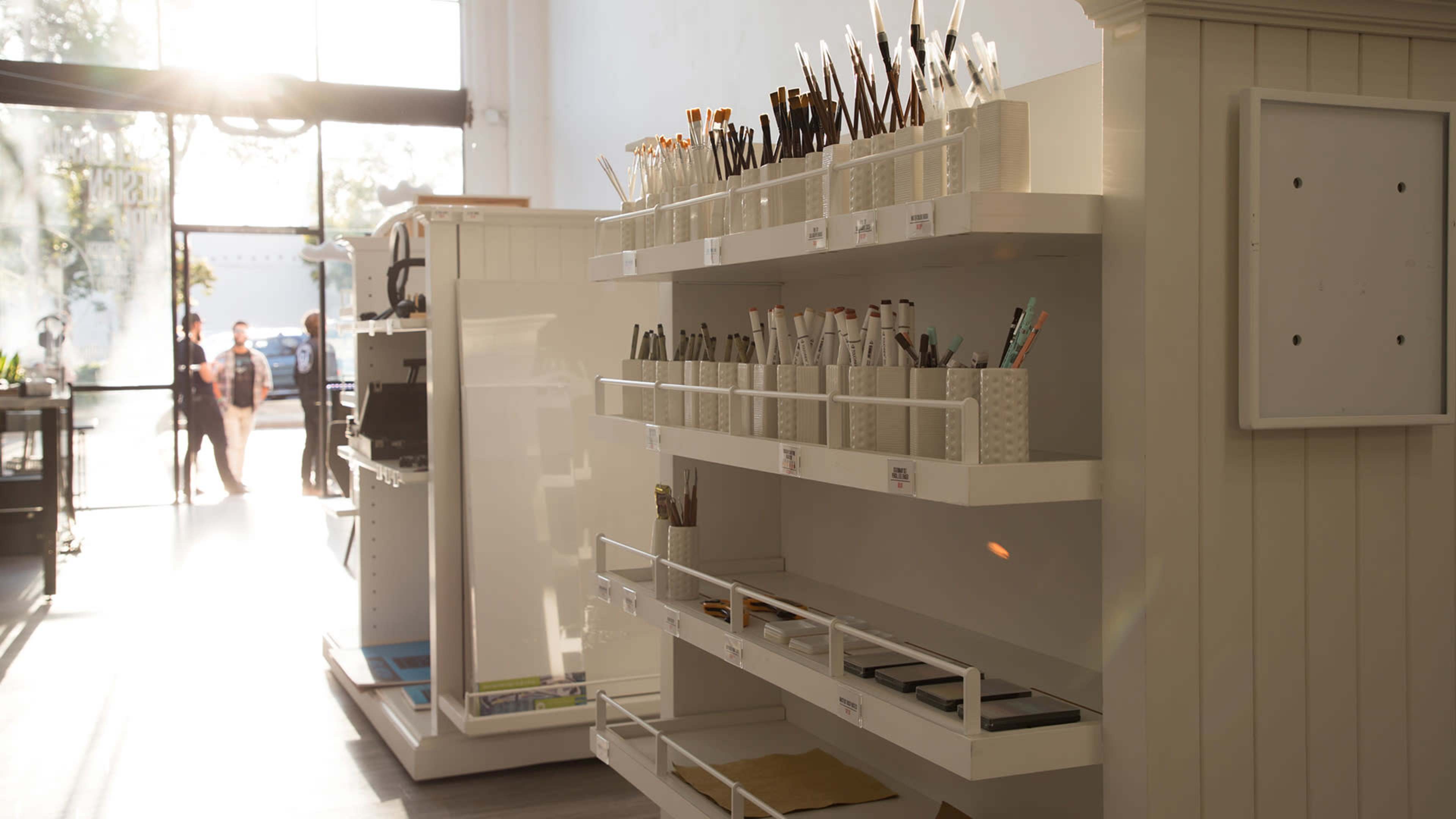 A well-organized store display with various pens and stationery items arranged on shelves, illuminated by natural light coming through the large front windows.