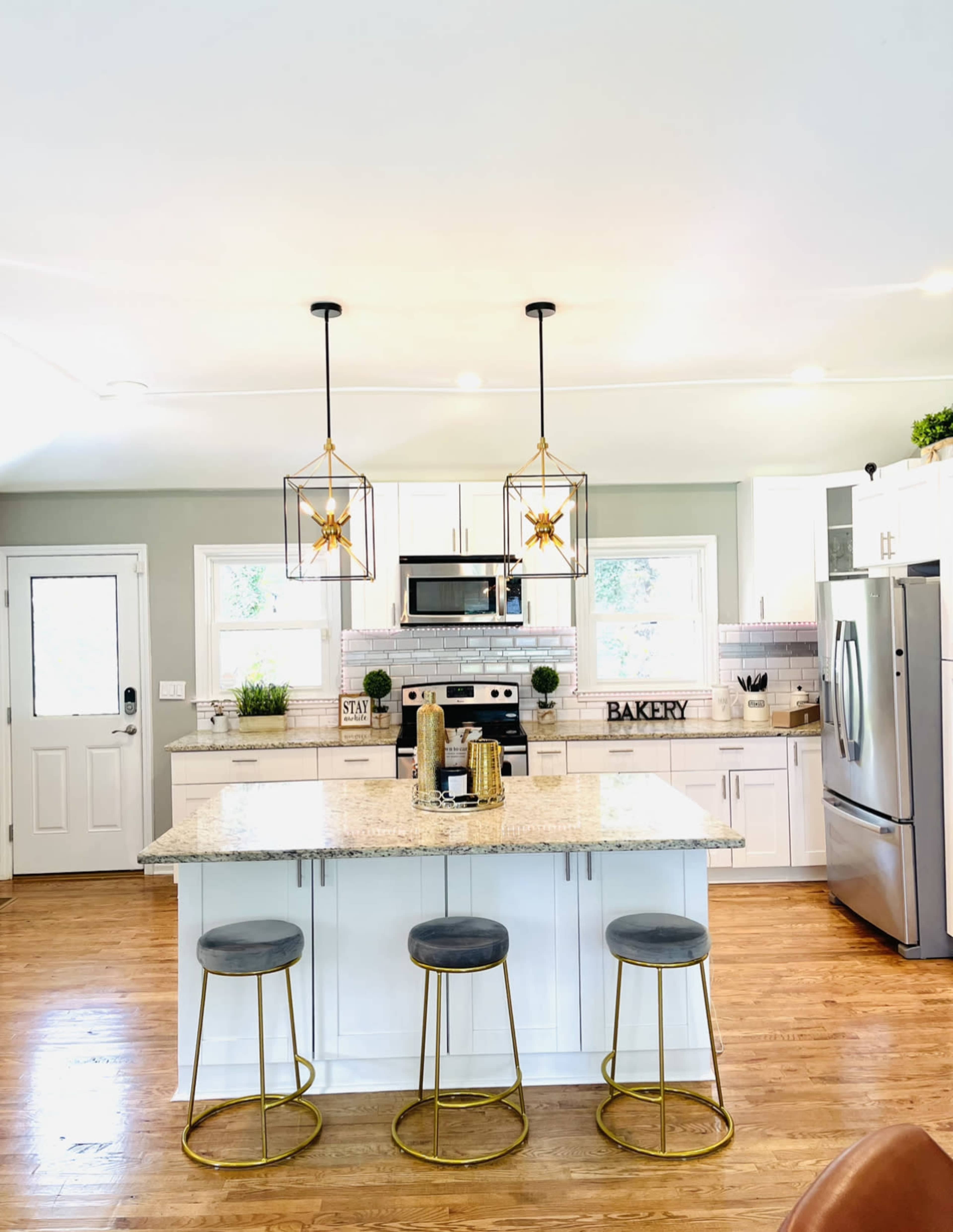 A modern kitchen features a large island with three gold-accented stools and two decorative pendant lights above.