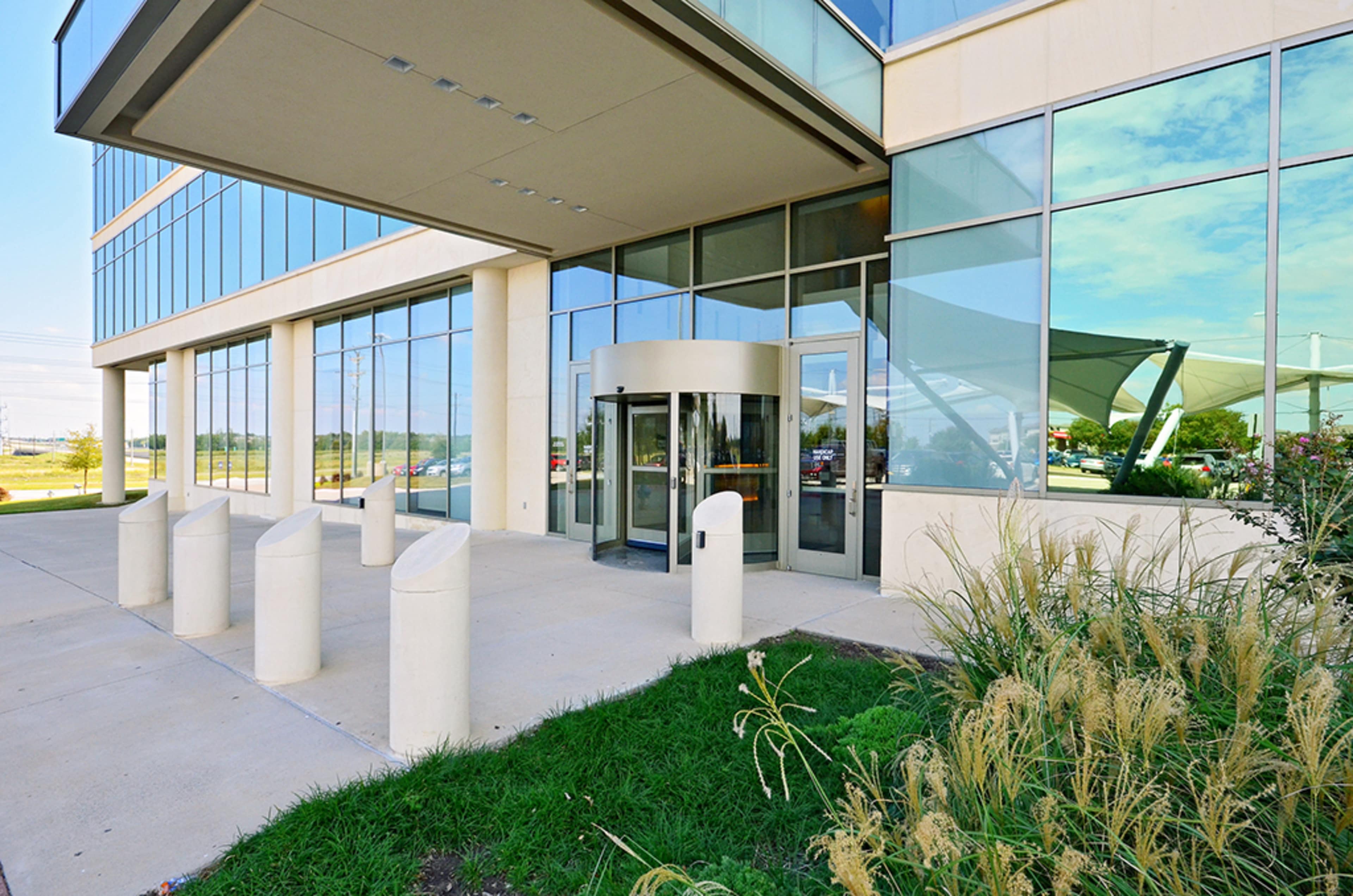 The entrance of a modern building featuring large glass windows, a revolving door, and white cylindrical bollards in front.