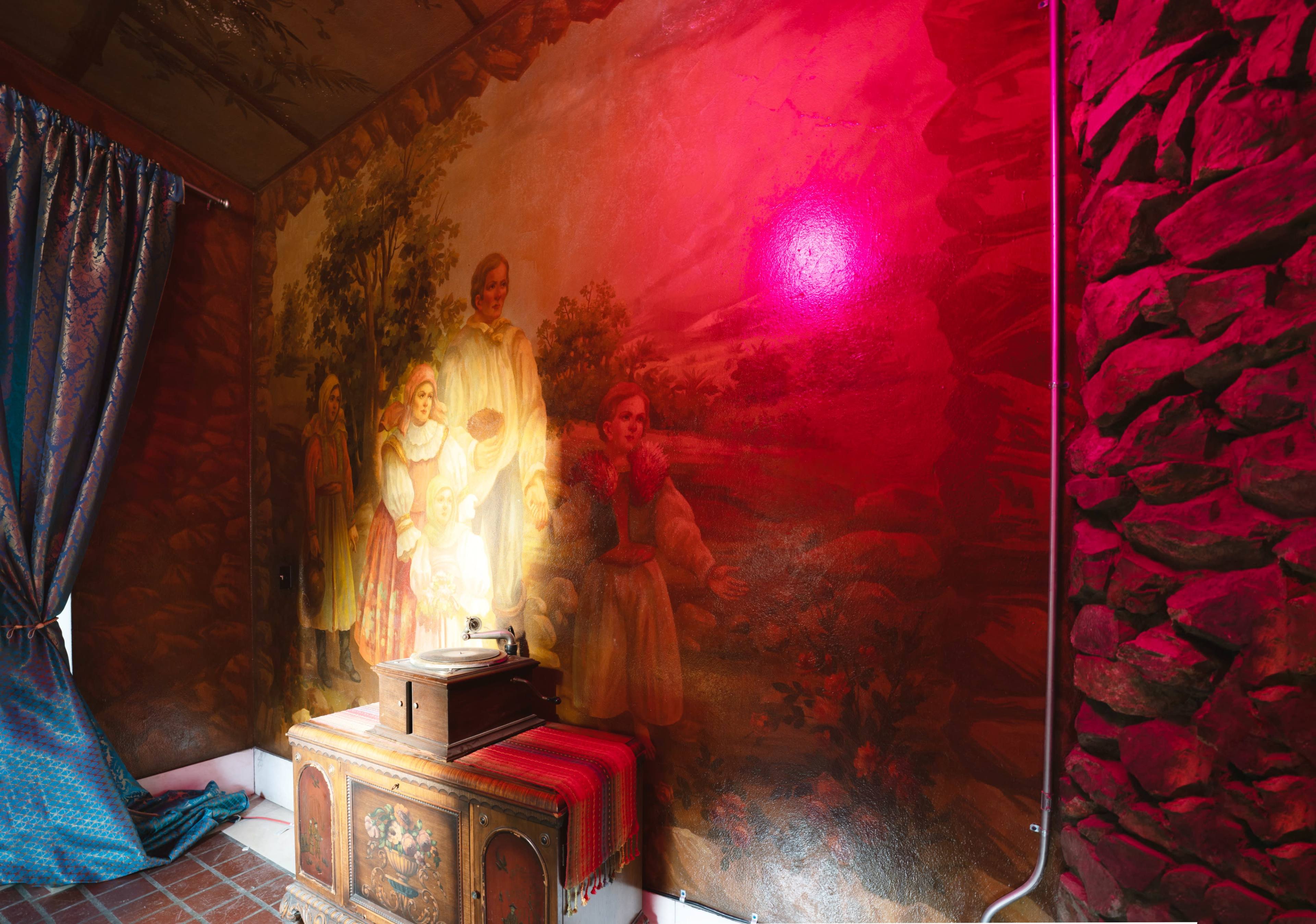 The image shows a vintage wooden cabinet with a gramophone, positioned against a wall featuring a large, colorful mural of people in a landscape, illuminated by a red light.