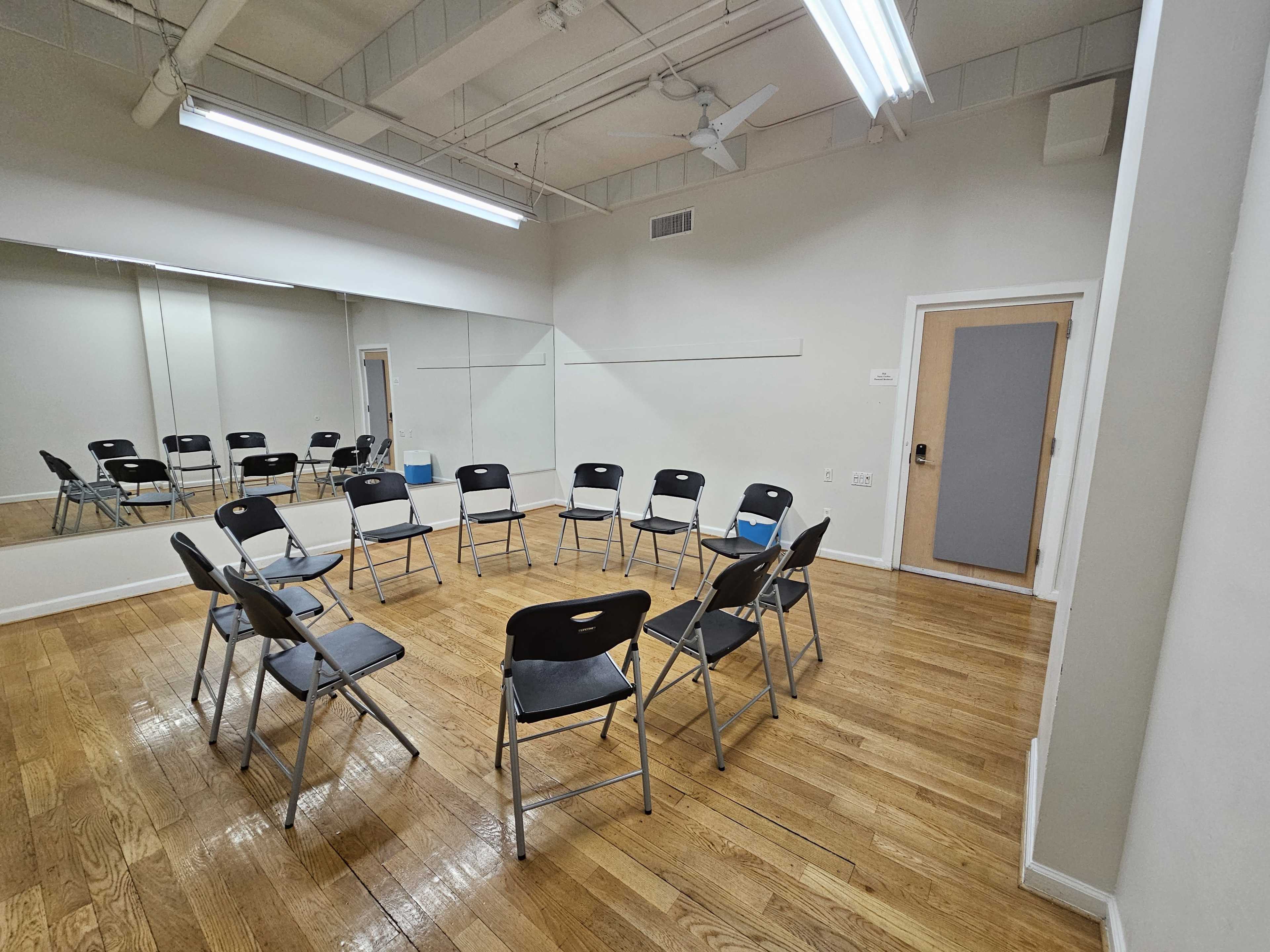 The image shows a room with wooden flooring, featuring a circle of black folding chairs and a large mirror on one wall.