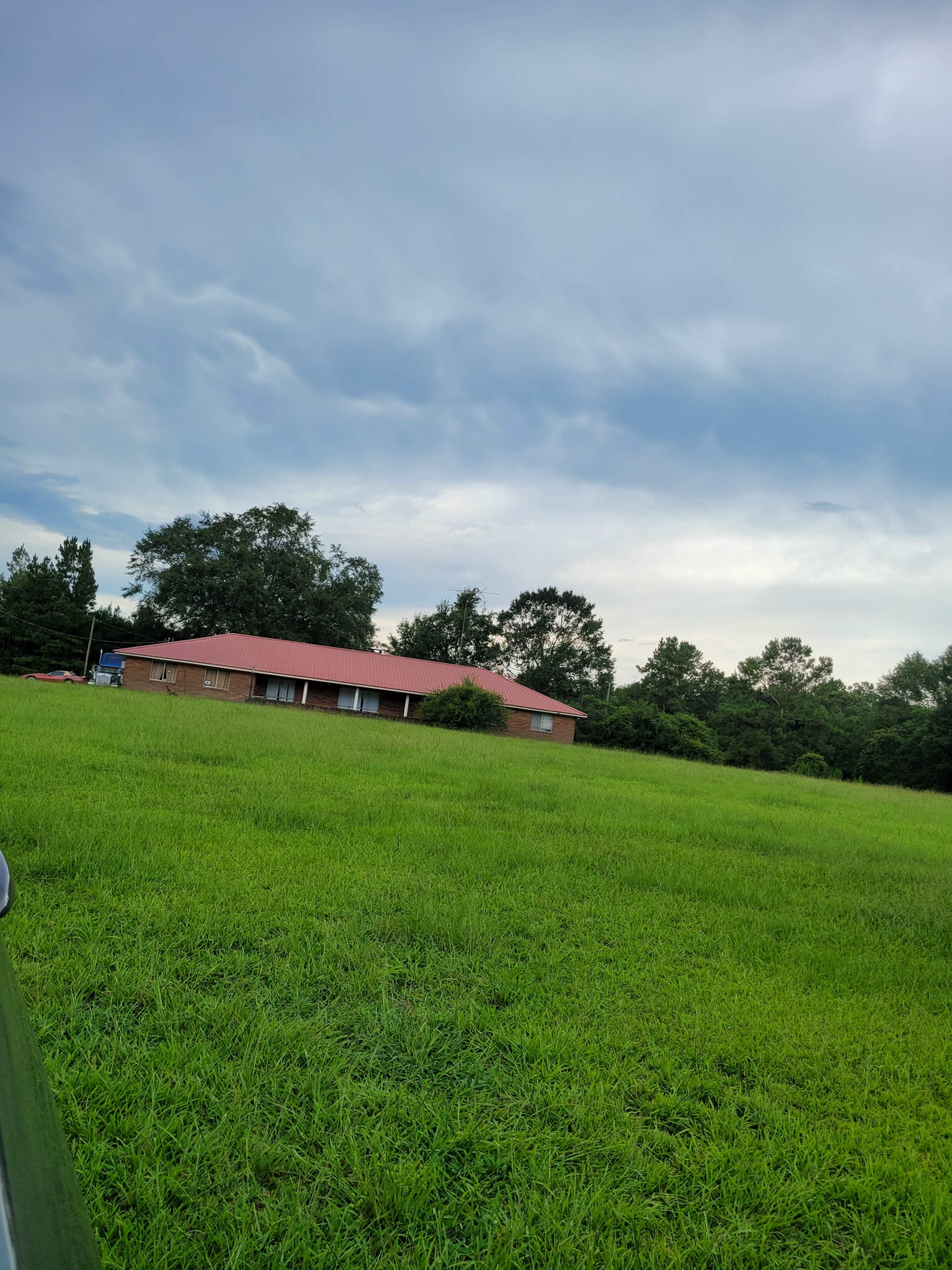 A single-story brick house with a red roof is situated next to a large green field under a cloudy sky.