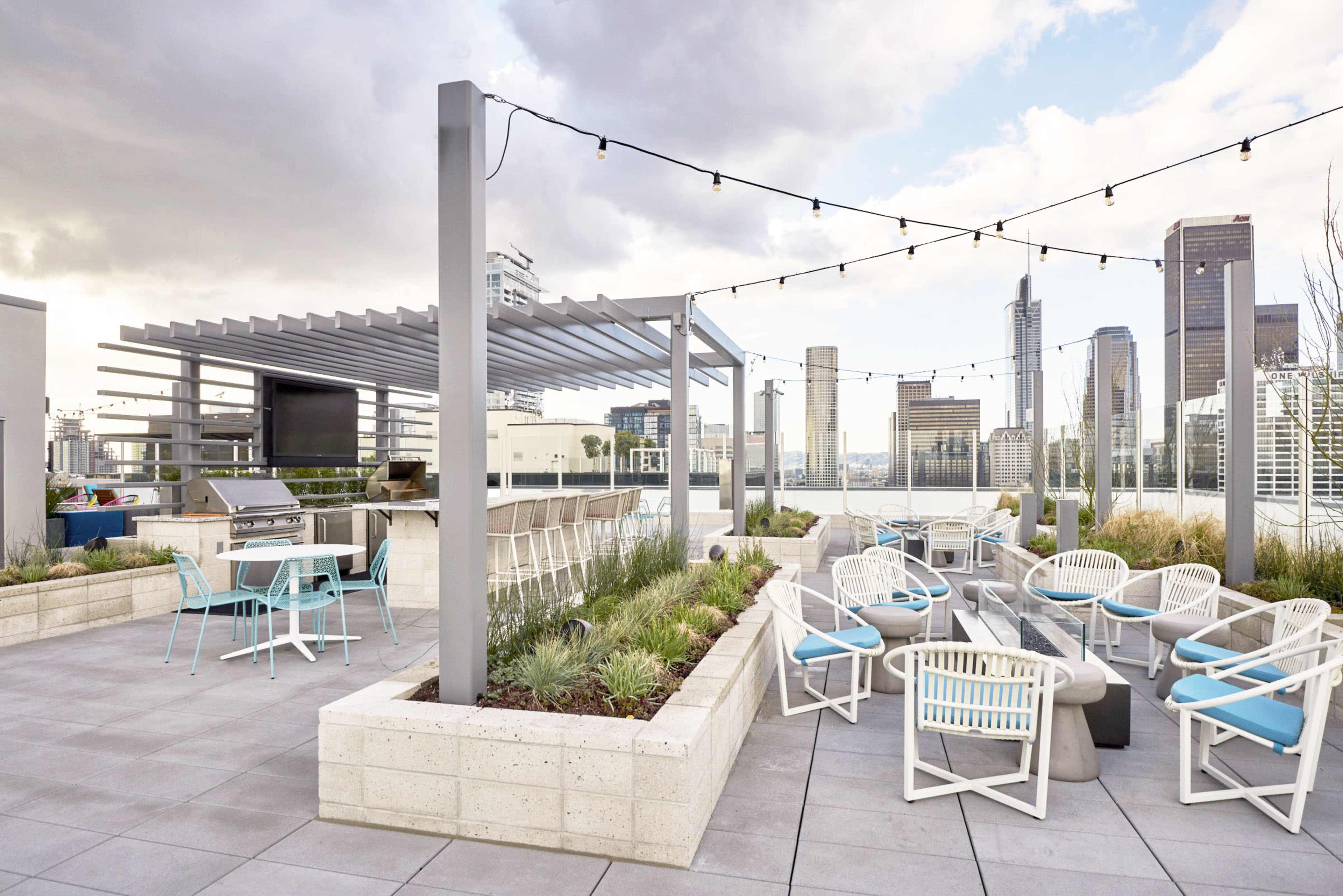 The image shows a rooftop terrace featuring outdoor seating, a barbecue area, and skyline views of tall buildings under a cloudy sky.