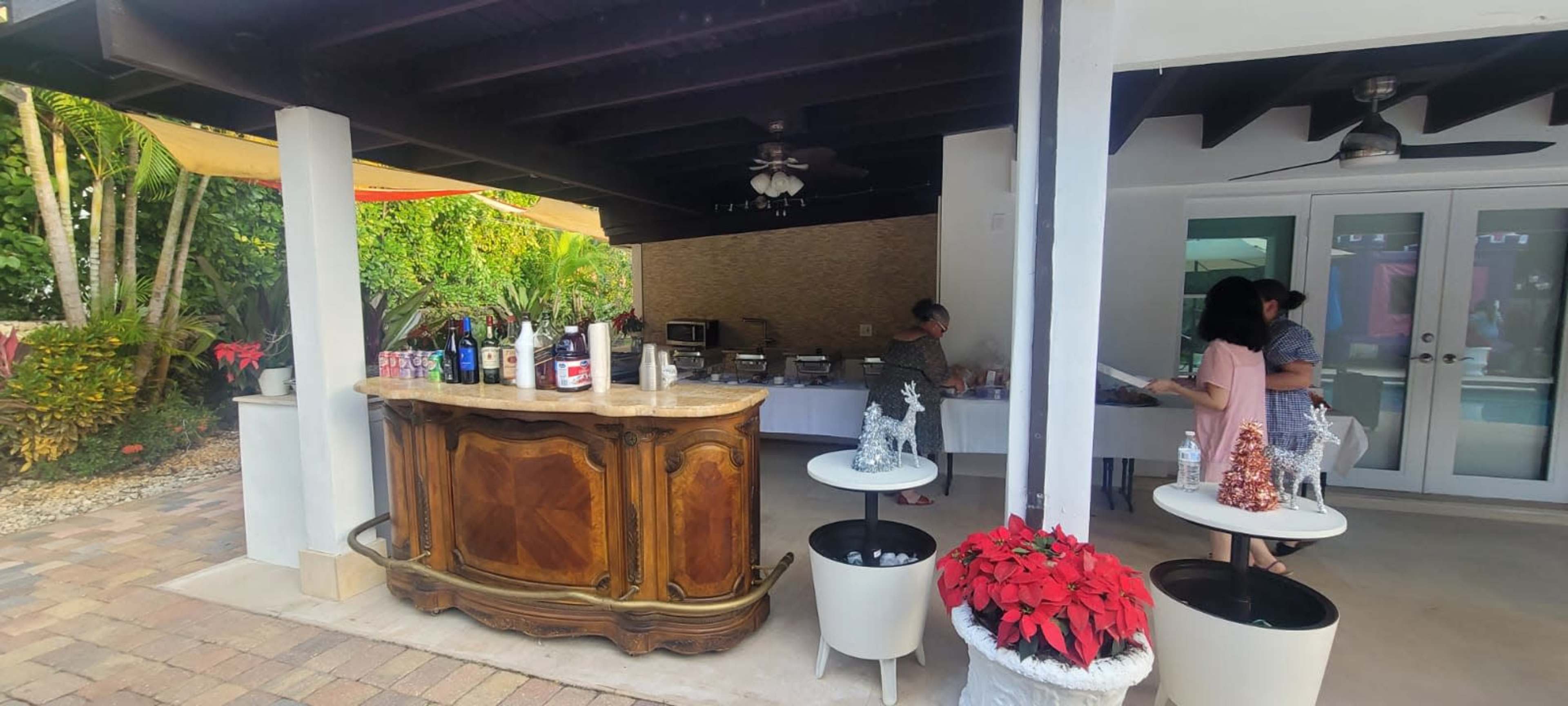 A bar area with a wooden counter is set up outdoors next to a buffet table, while two people are conversing nearby in a tropical garden setting.
