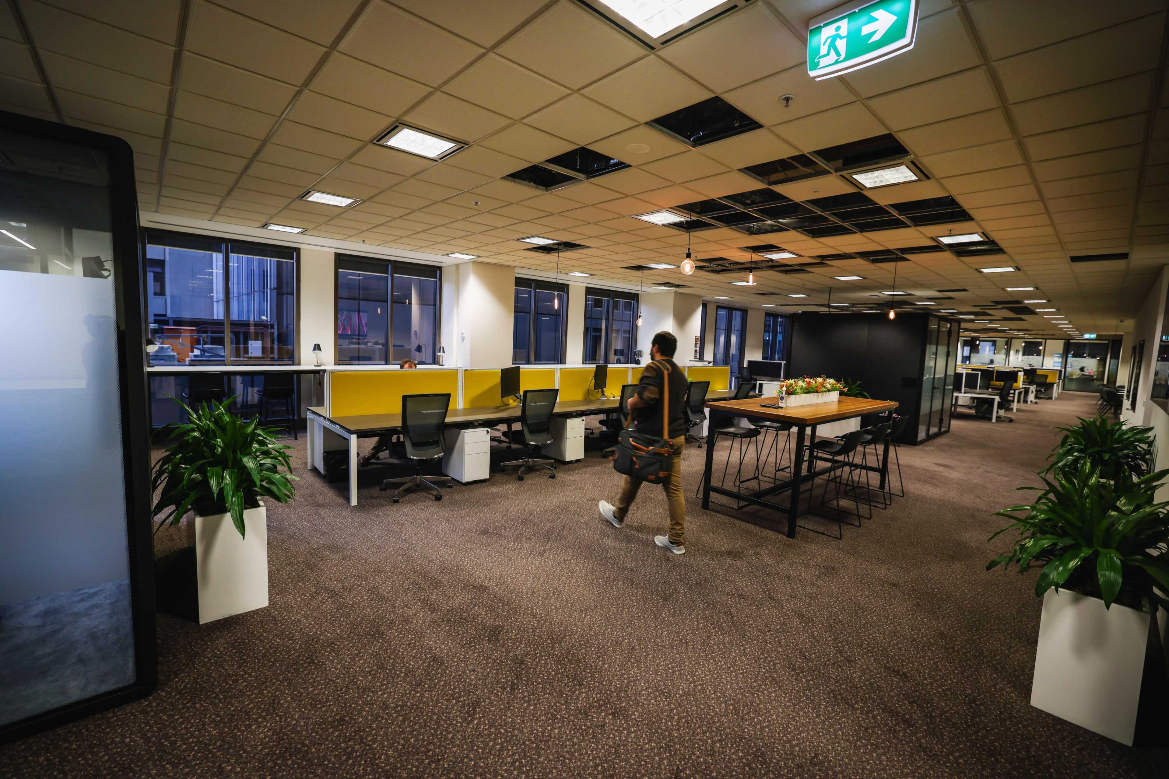 A person walks through a spacious office environment featuring multiple workstations, plants, and a large communal table.