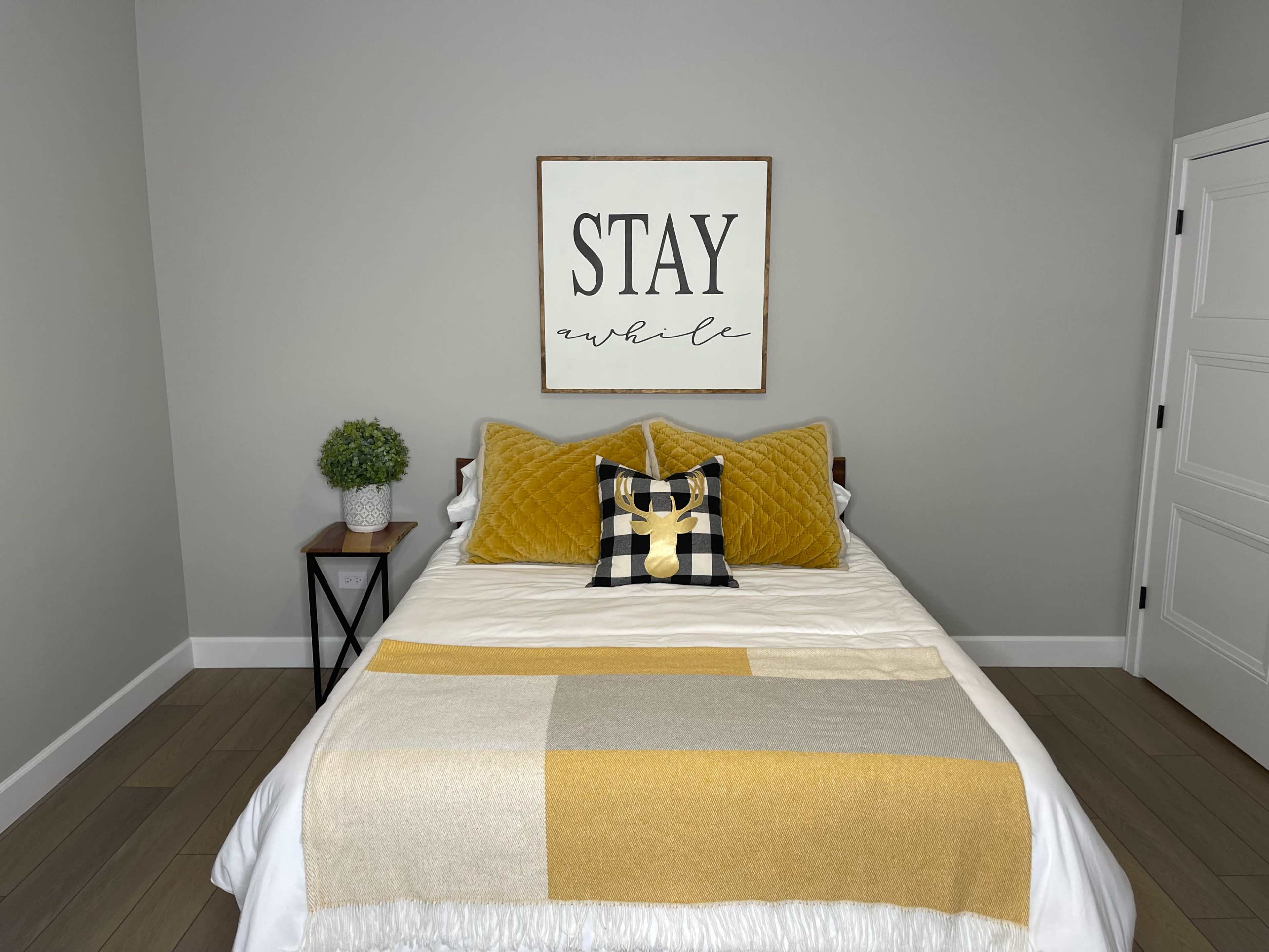 The image shows a neatly made bed with yellow and black decorative pillows, a light-colored throw blanket, and a wall art piece that reads "STAY awhile" above it, set in a simple bedroom with gray walls.