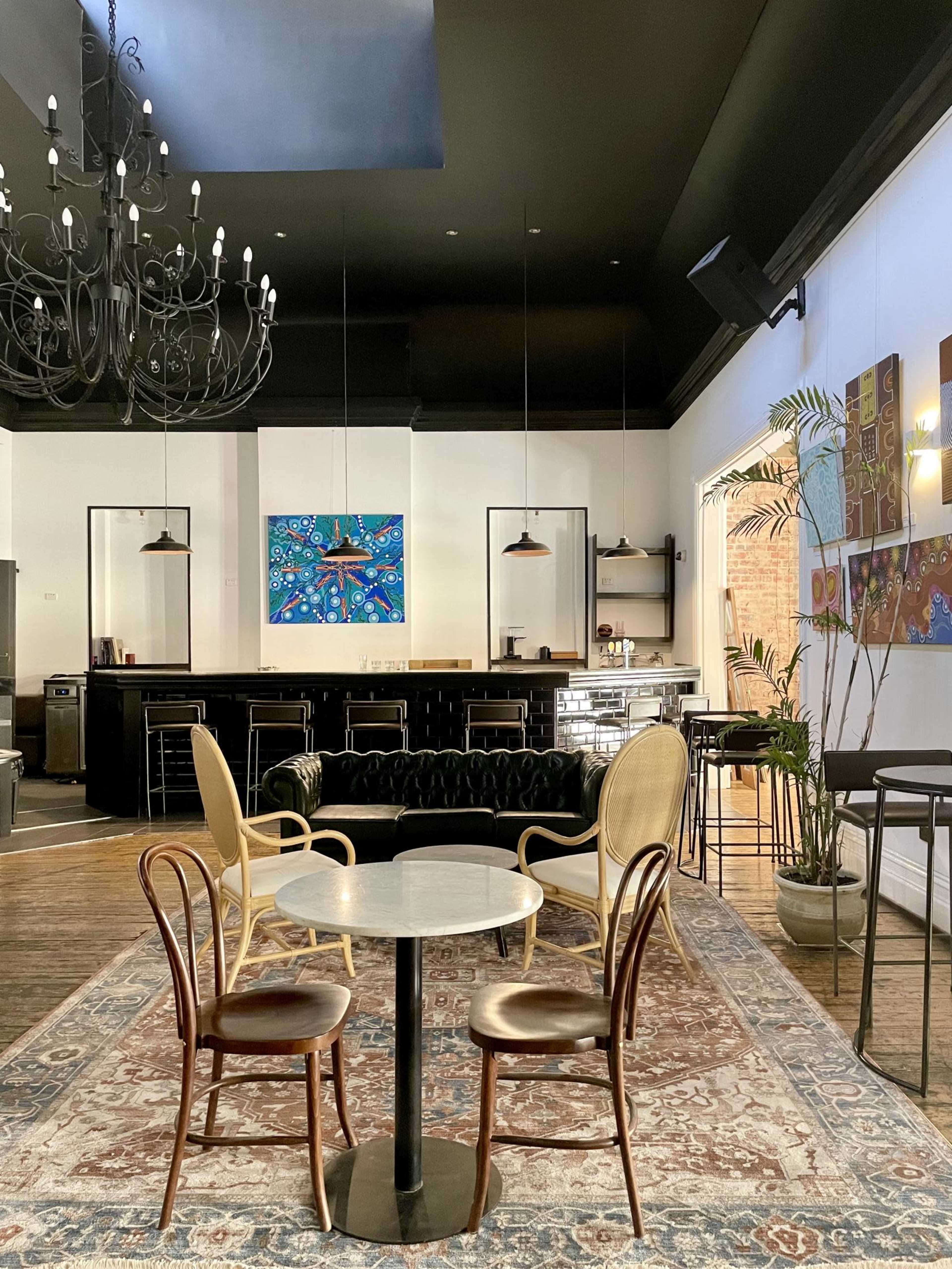 A modern cafe interior featuring a round marble table surrounded by three chairs, with a bar area and an artistic painting on the wall.