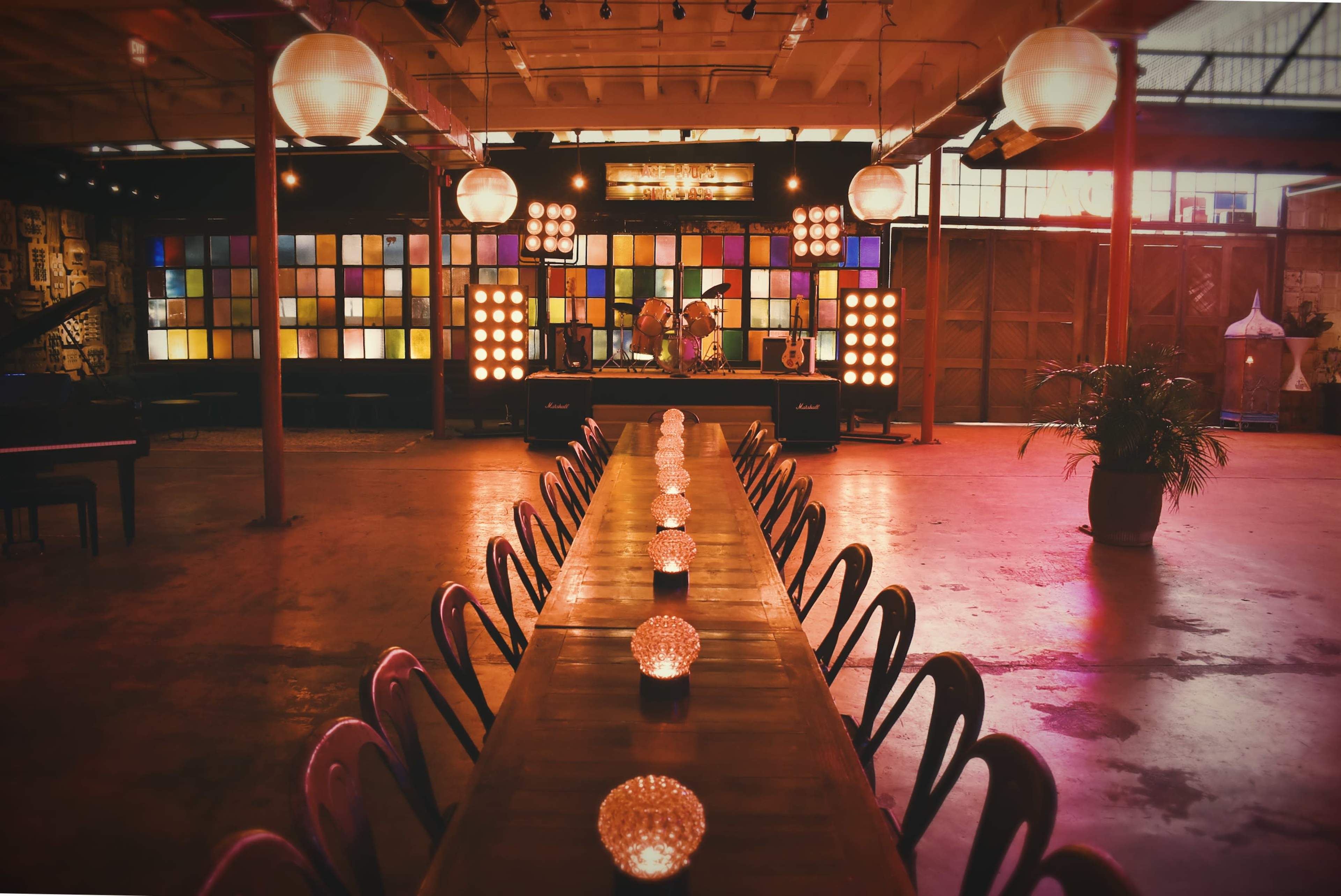 The image depicts a spacious interior of an event venue with a long wooden table set with candle holders, a DJ booth in the background, and colorful stained glass windows lining the walls.