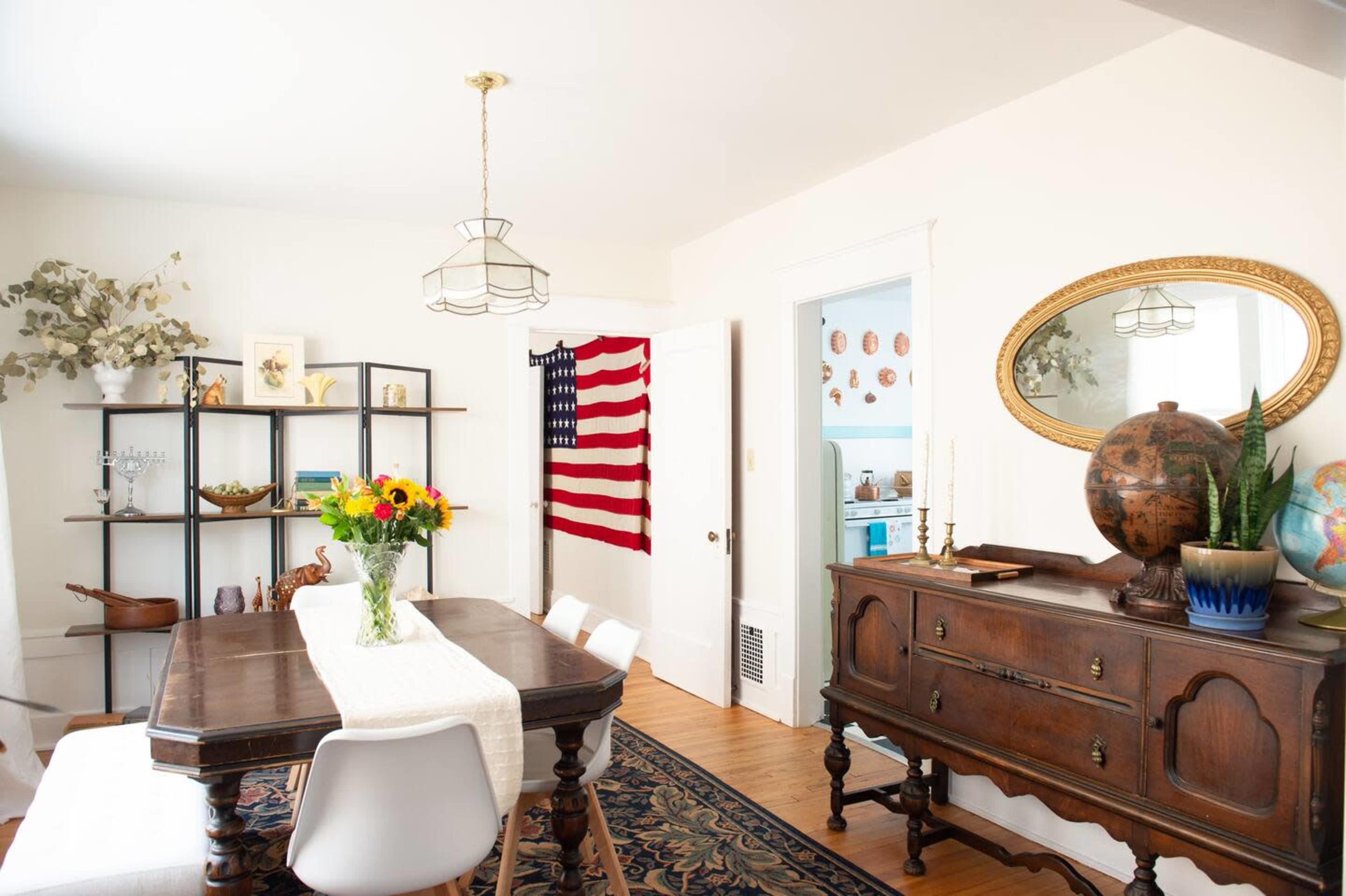 A dining room features a wooden table surrounded by white chairs, with an American flag hanging on the wall and a globe on the sideboard.