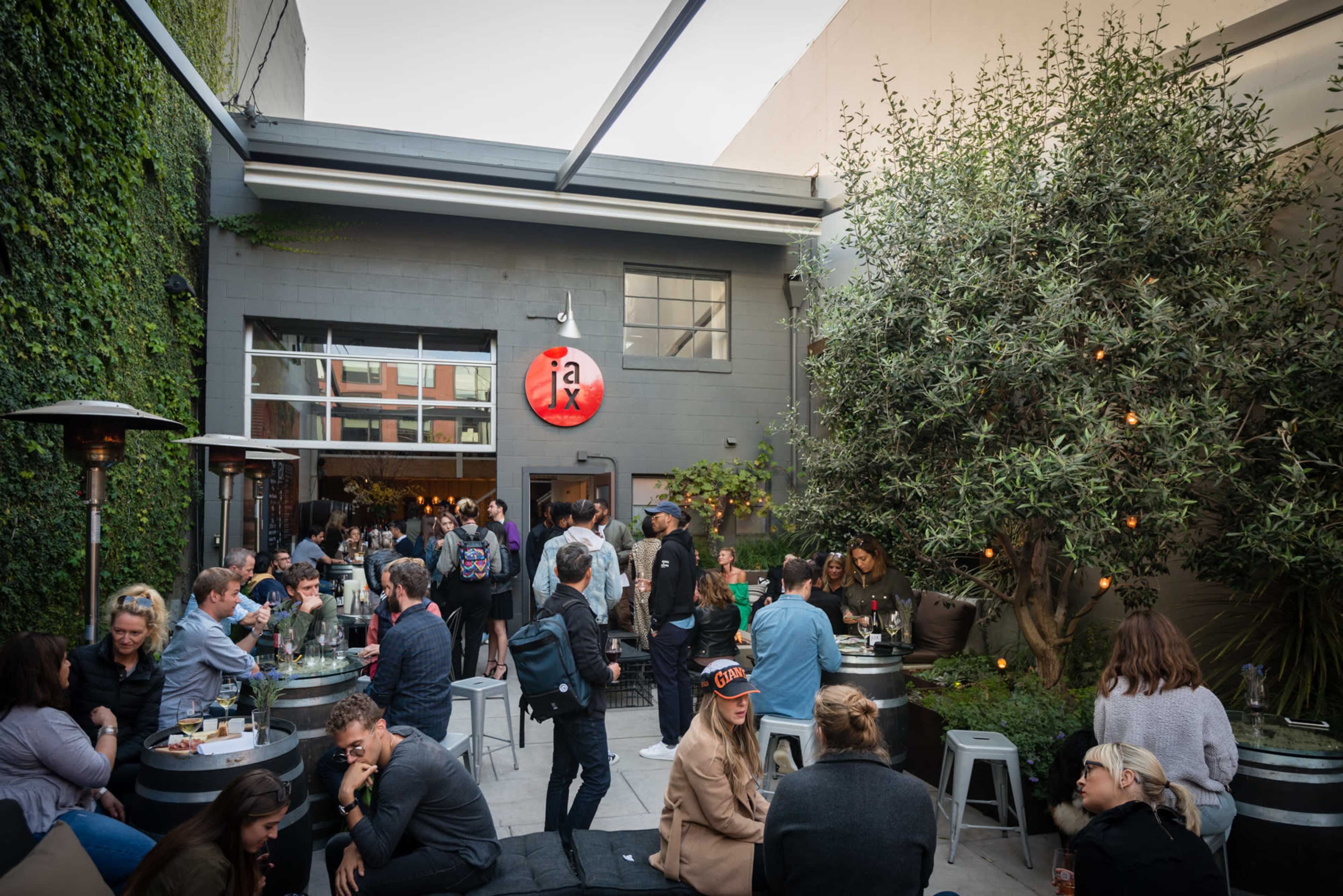 A bustling outdoor courtyard filled with people socializing around tables, with a building featuring a red circular sign that reads "ja."