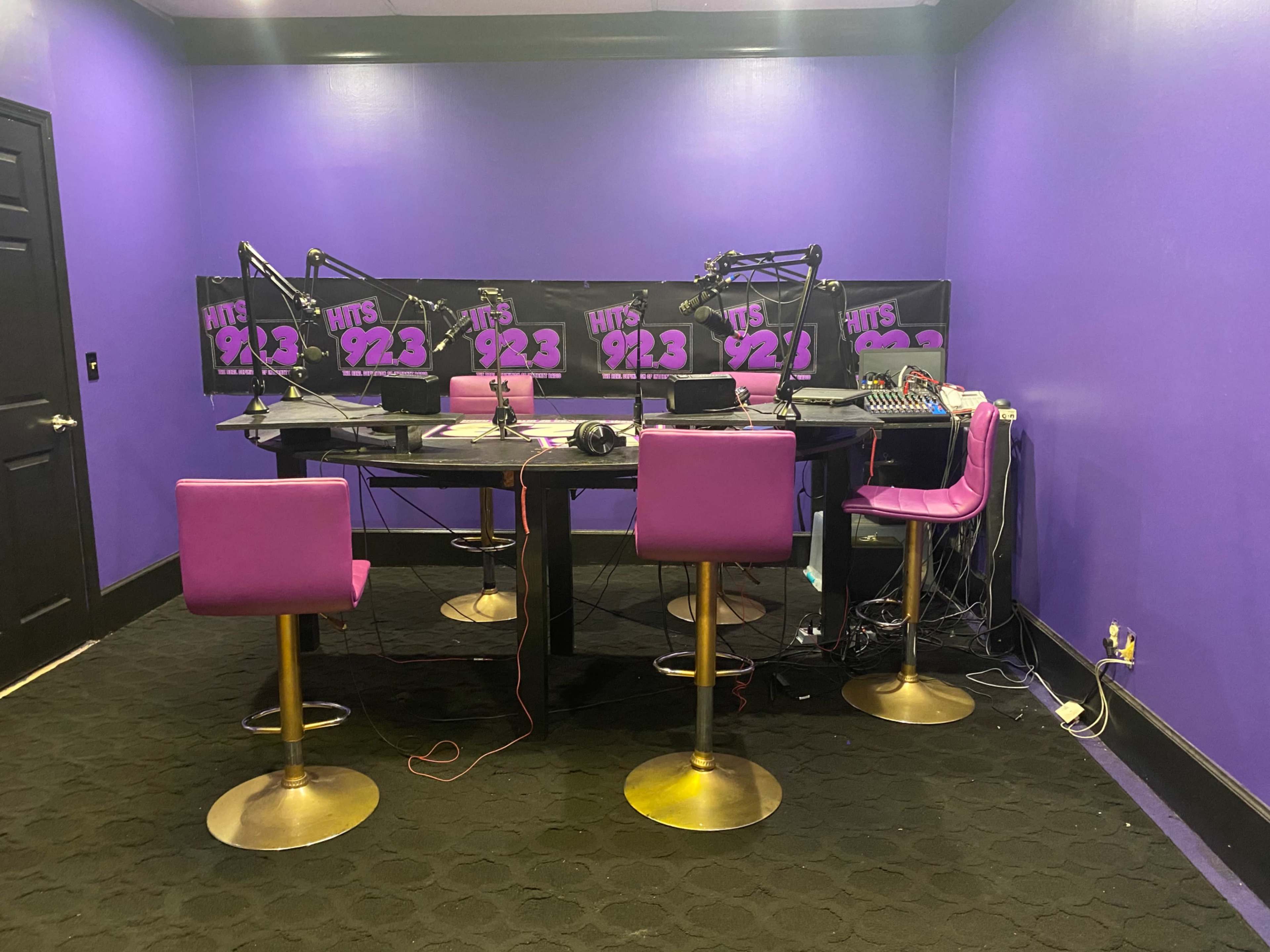 A radio studio with a black table, purple walls, and pink barstools.