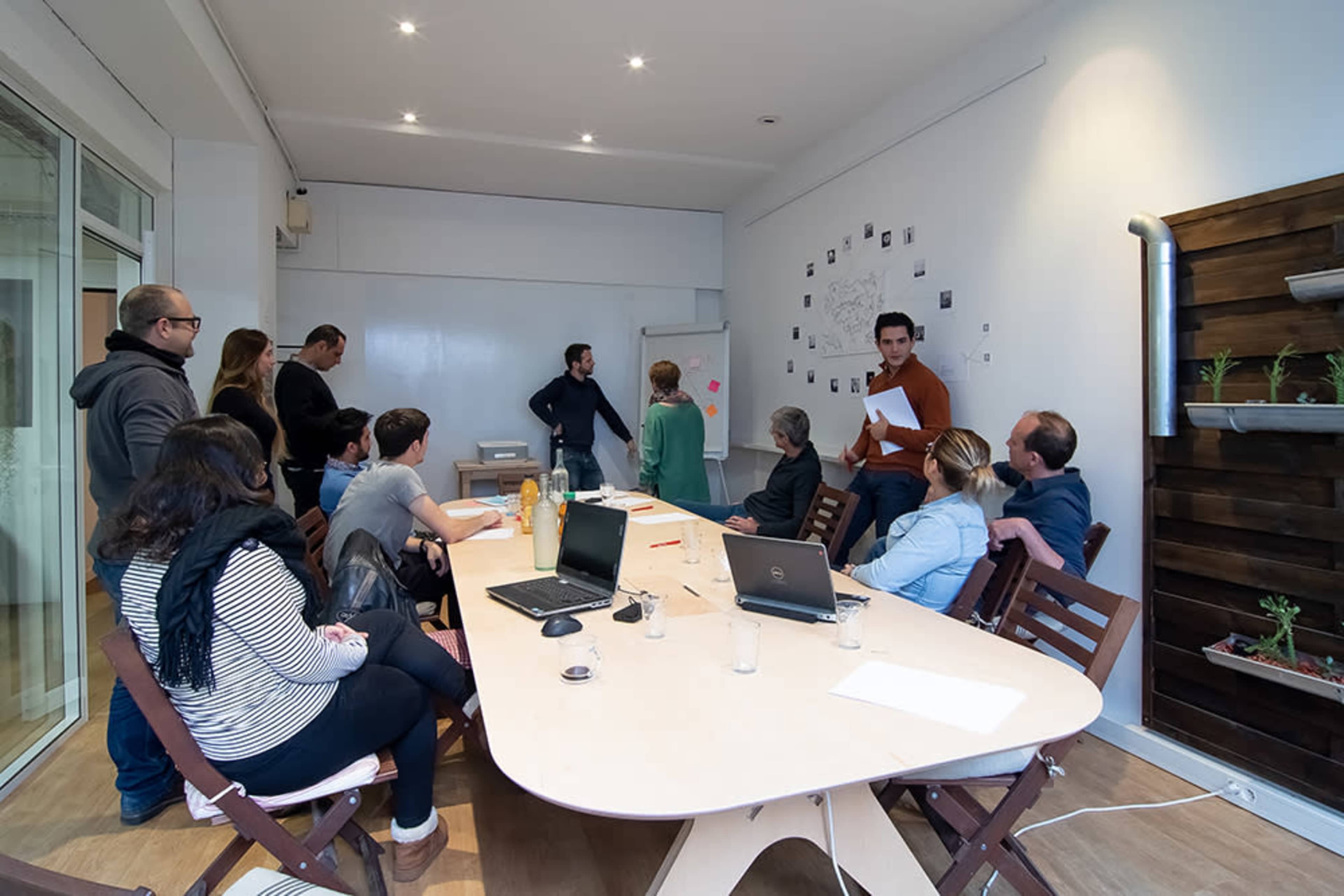 A group of ten people engages in a discussion around a large table in a meeting room with a whiteboard and wall-mounted plants.