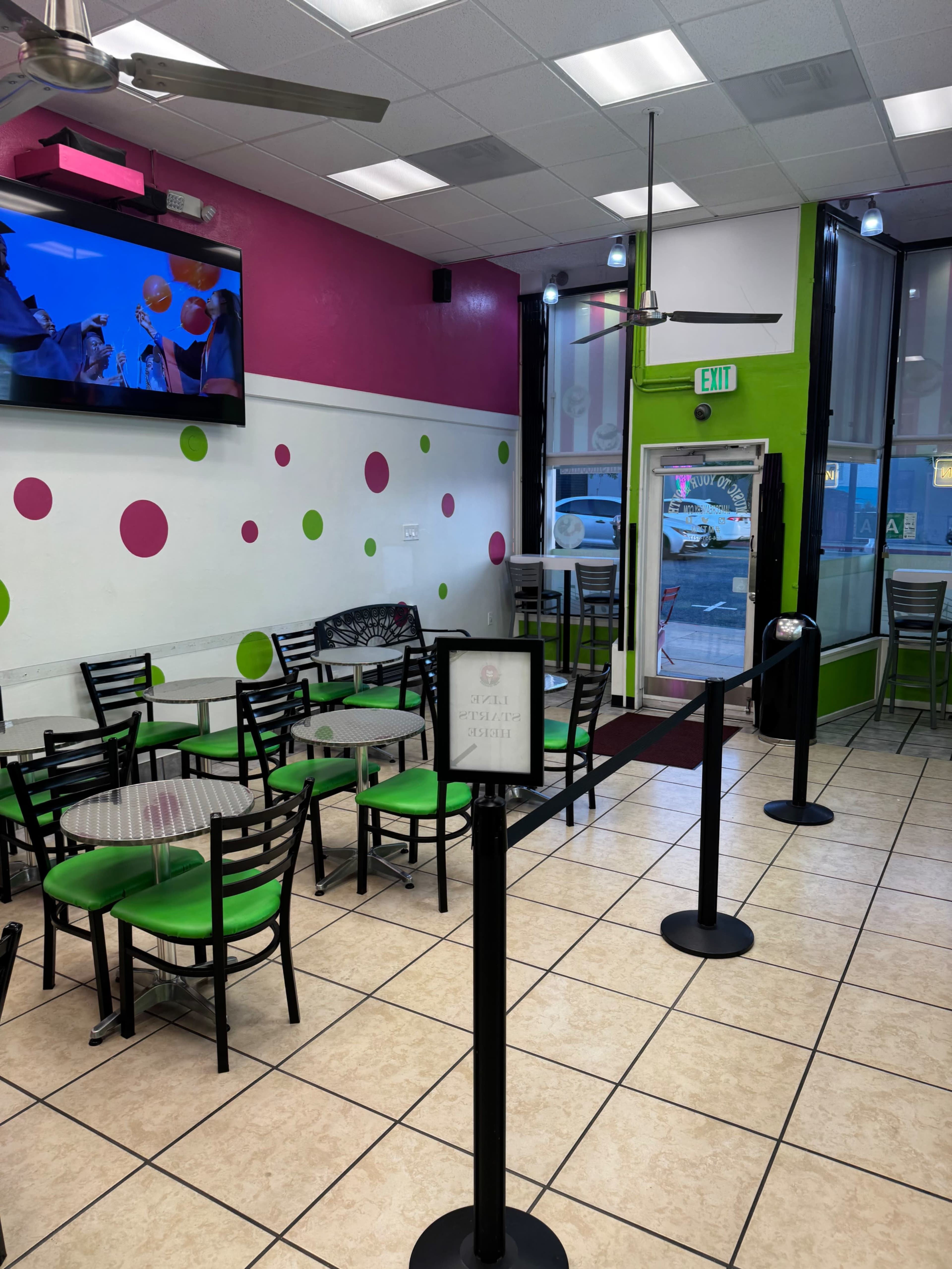 The image shows a brightly colored cafe with green and pink decor, featuring several tables and chairs arranged neatly, and a large TV mounted on the wall.