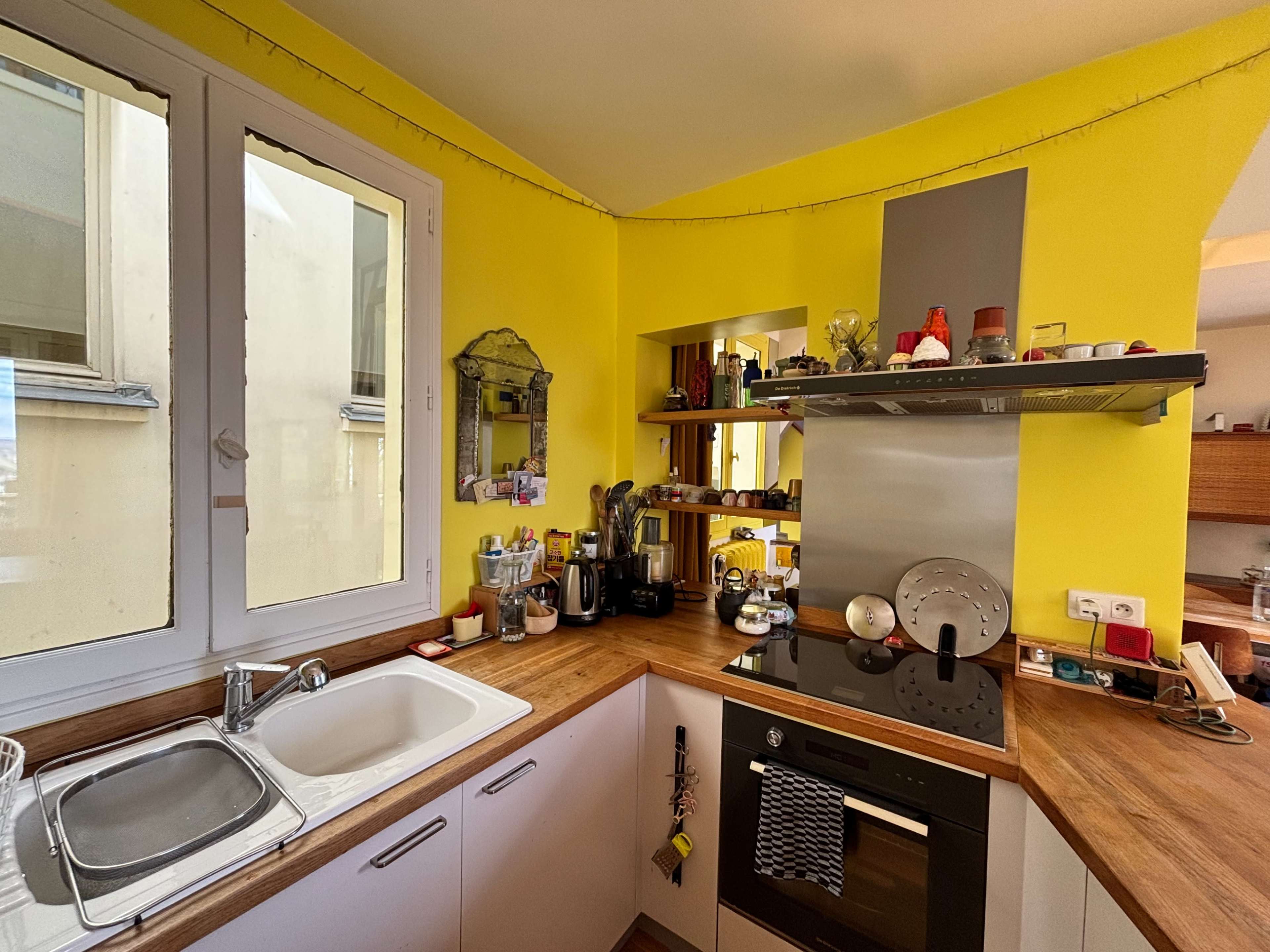 The image shows a compact kitchen with yellow walls, modern appliances, and wooden countertops, featuring a sink and various kitchen items on shelves.