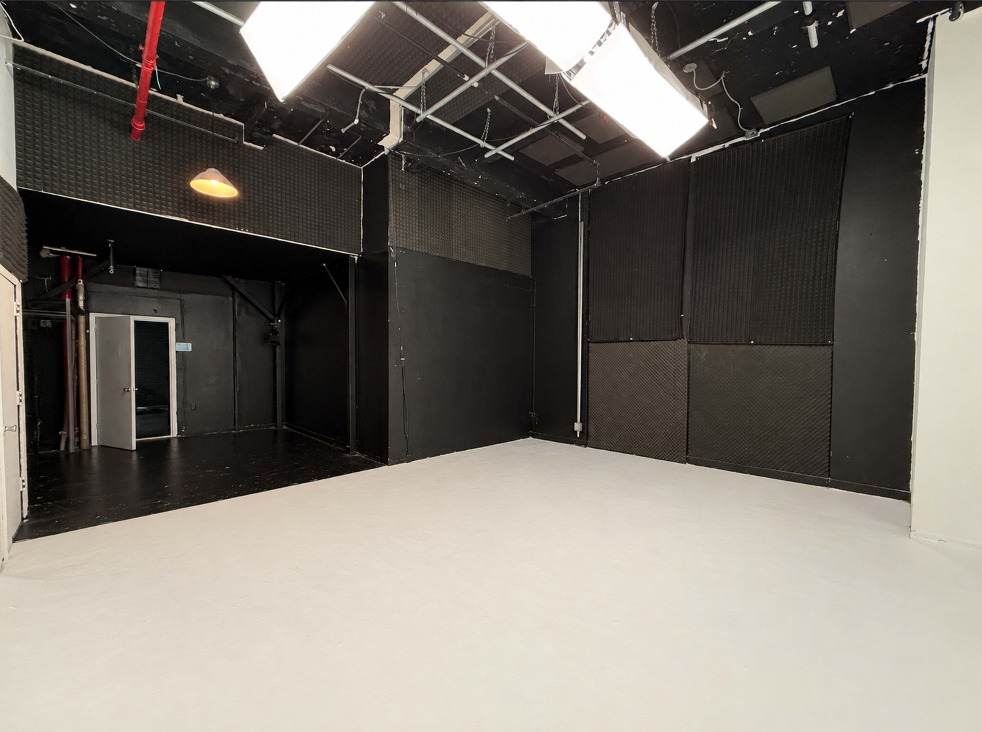 The image shows an empty studio space with black walls, a white floor, and overhead lighting.
