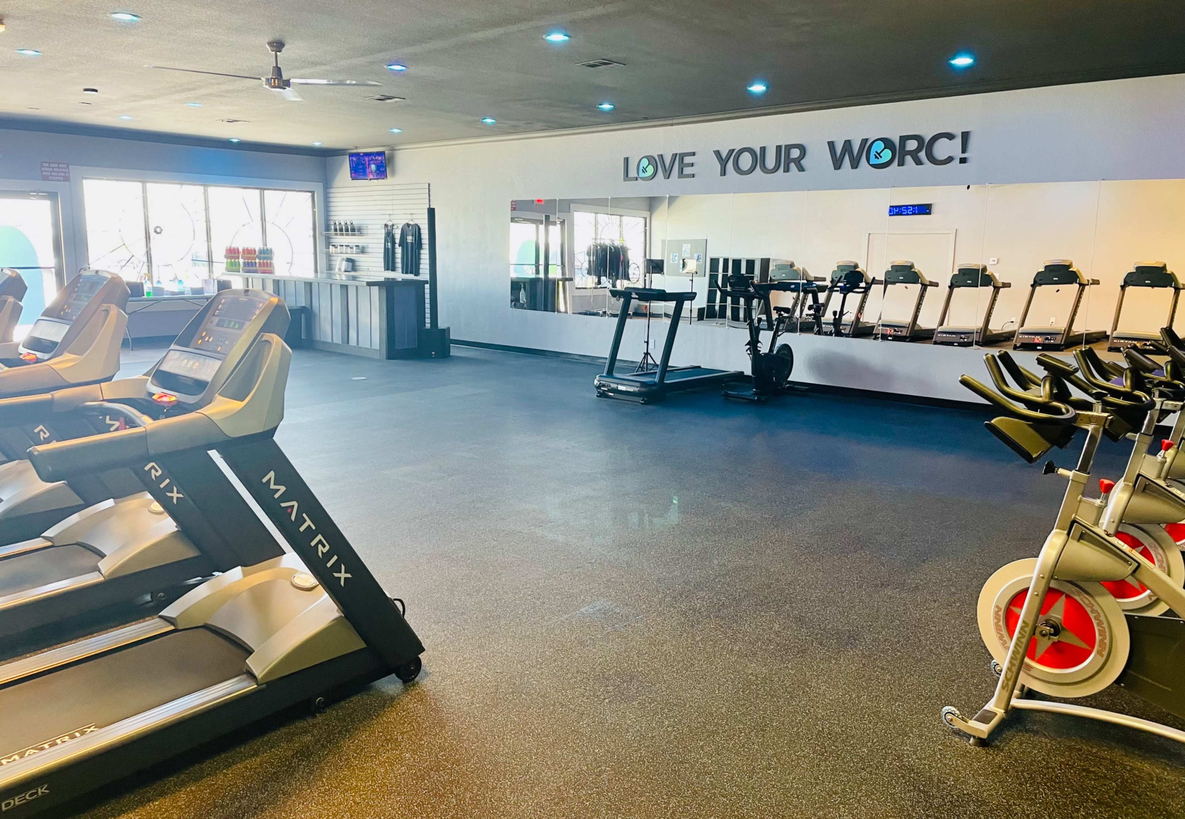 The image shows a gym interior featuring treadmills, stationary bikes, and a mirrored wall with the phrase "LOVE YOUR WORC!" displayed.