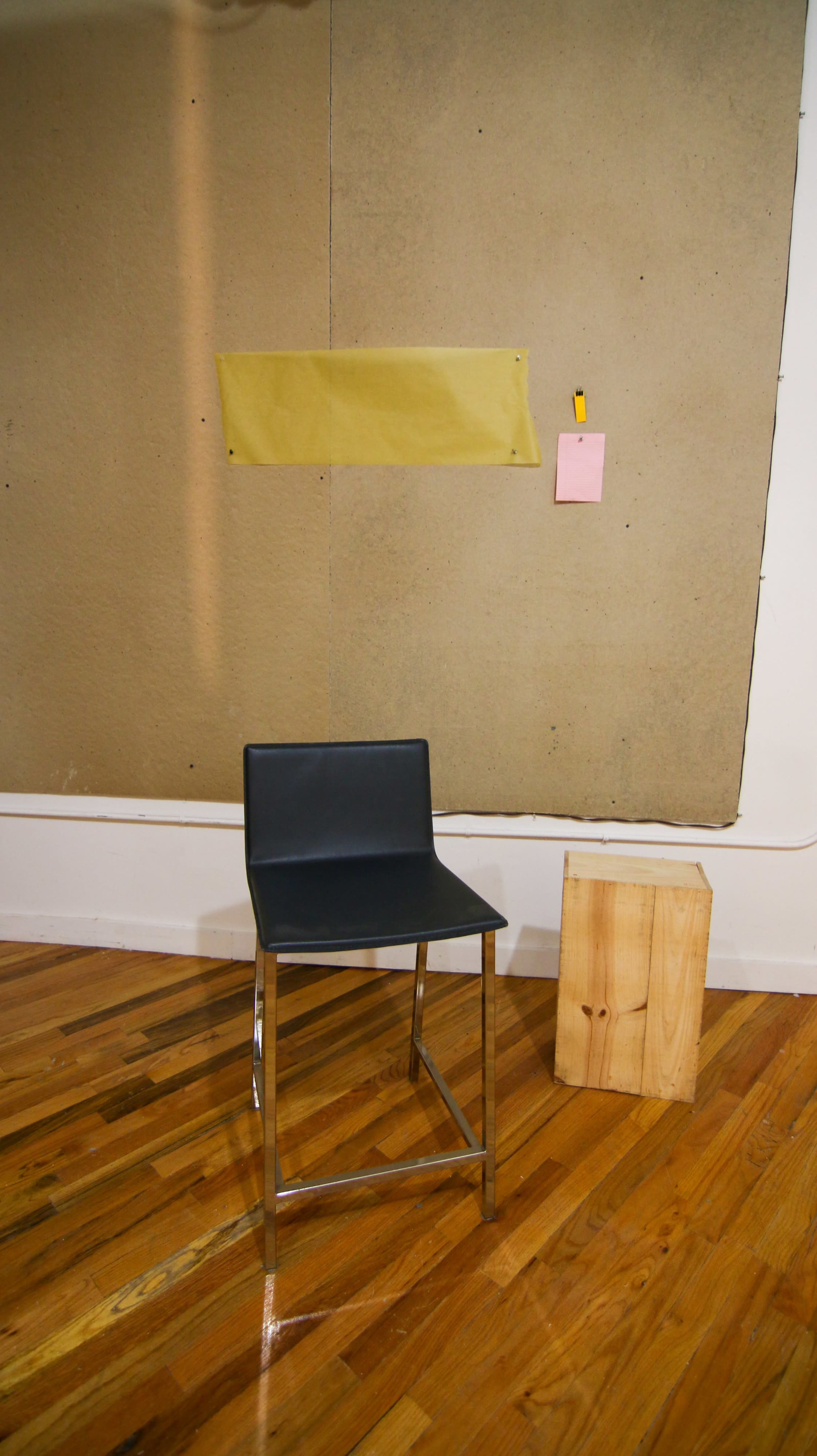A black metal-framed chair stands in front of a large, beige textured board mounted on the wall, with a wooden block on the floor beside it.