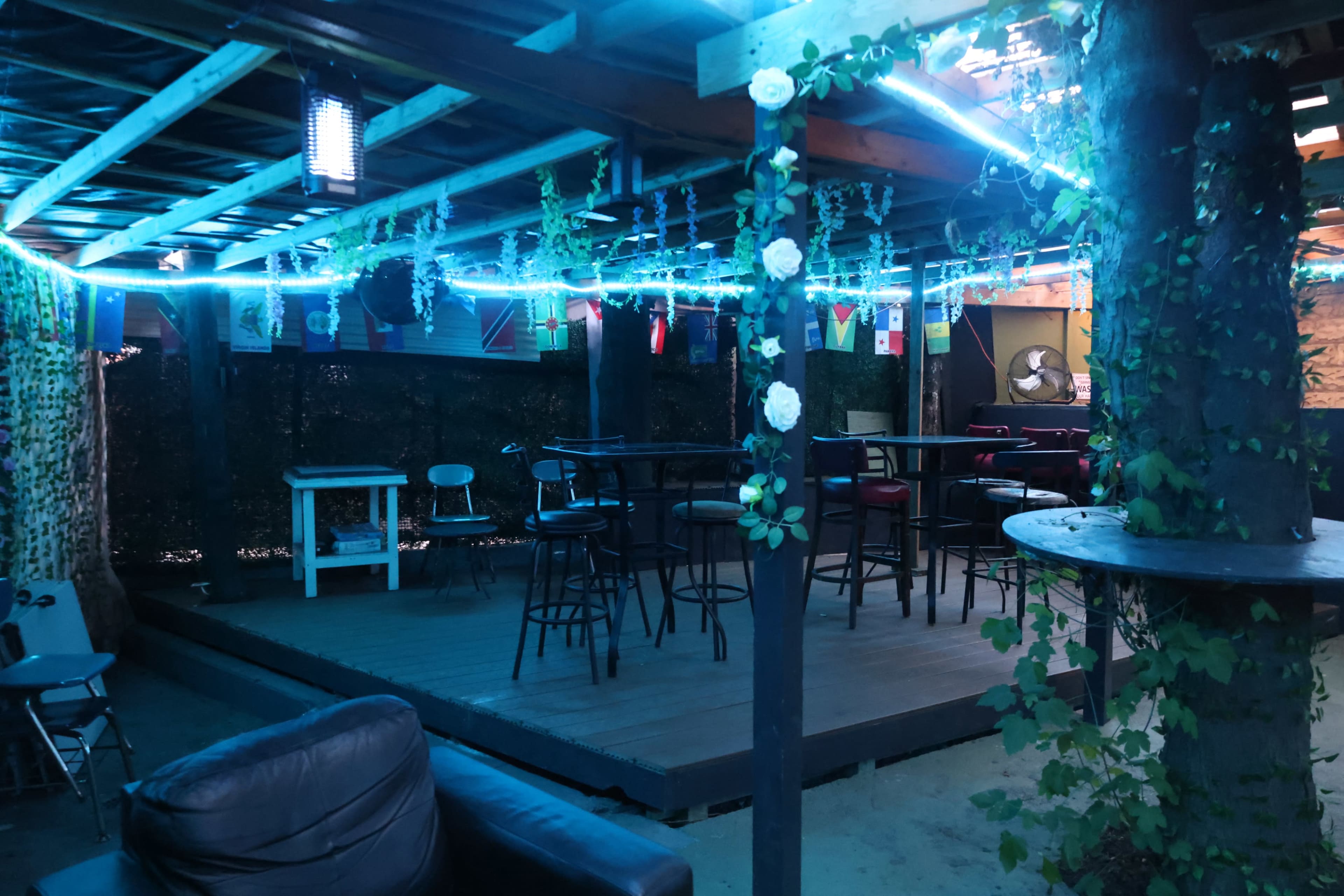 The image shows a dimly lit outdoor space featuring a bar area with several high stools, surrounded by decorative lights and greenery.