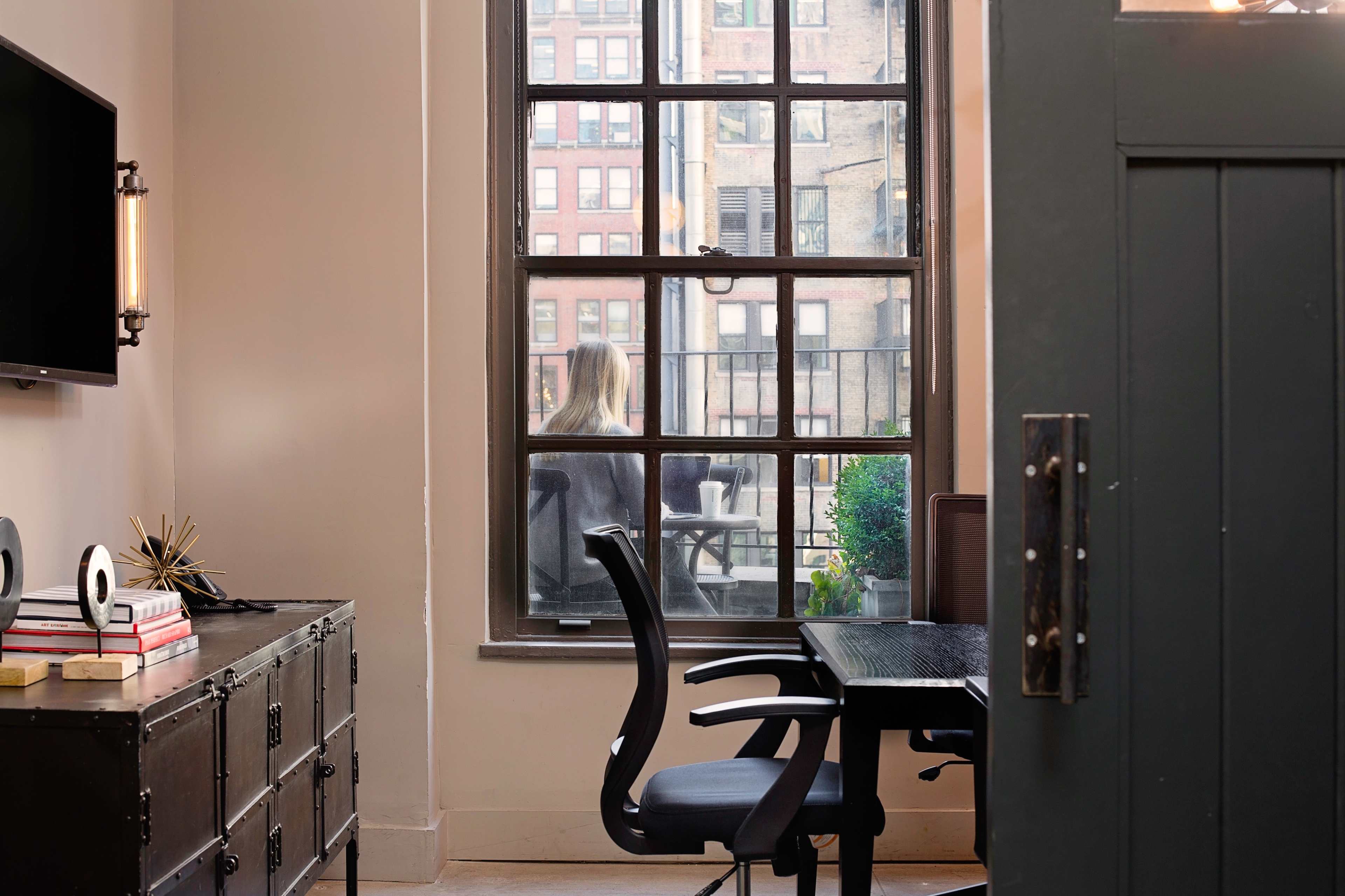 A modern office space features a desk and chair near a window overlooking an outdoor terrace with a person visible in the background.