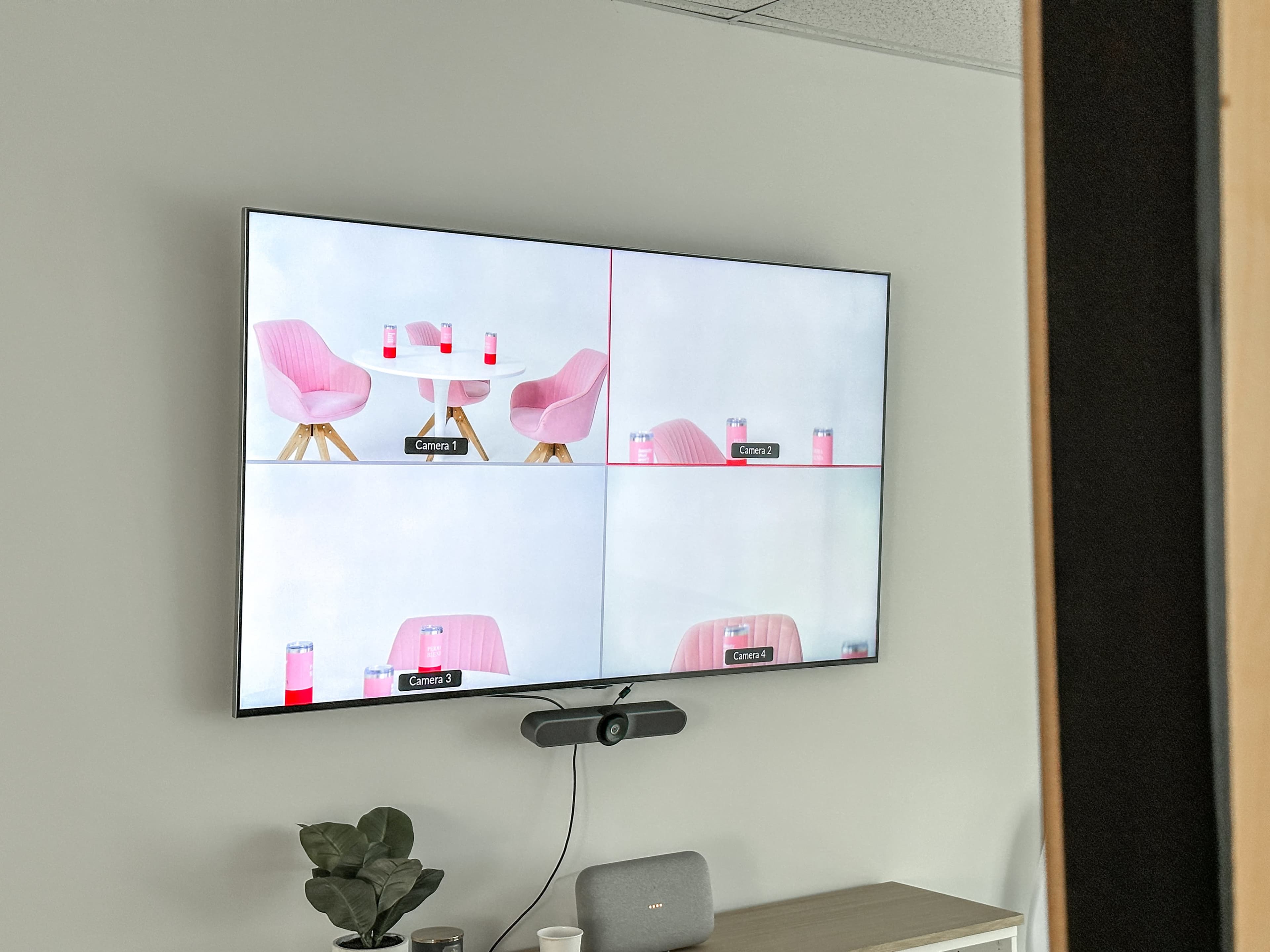 A large television displays a four-panel view of two pink chairs and drinks on small tables from different camera angles.