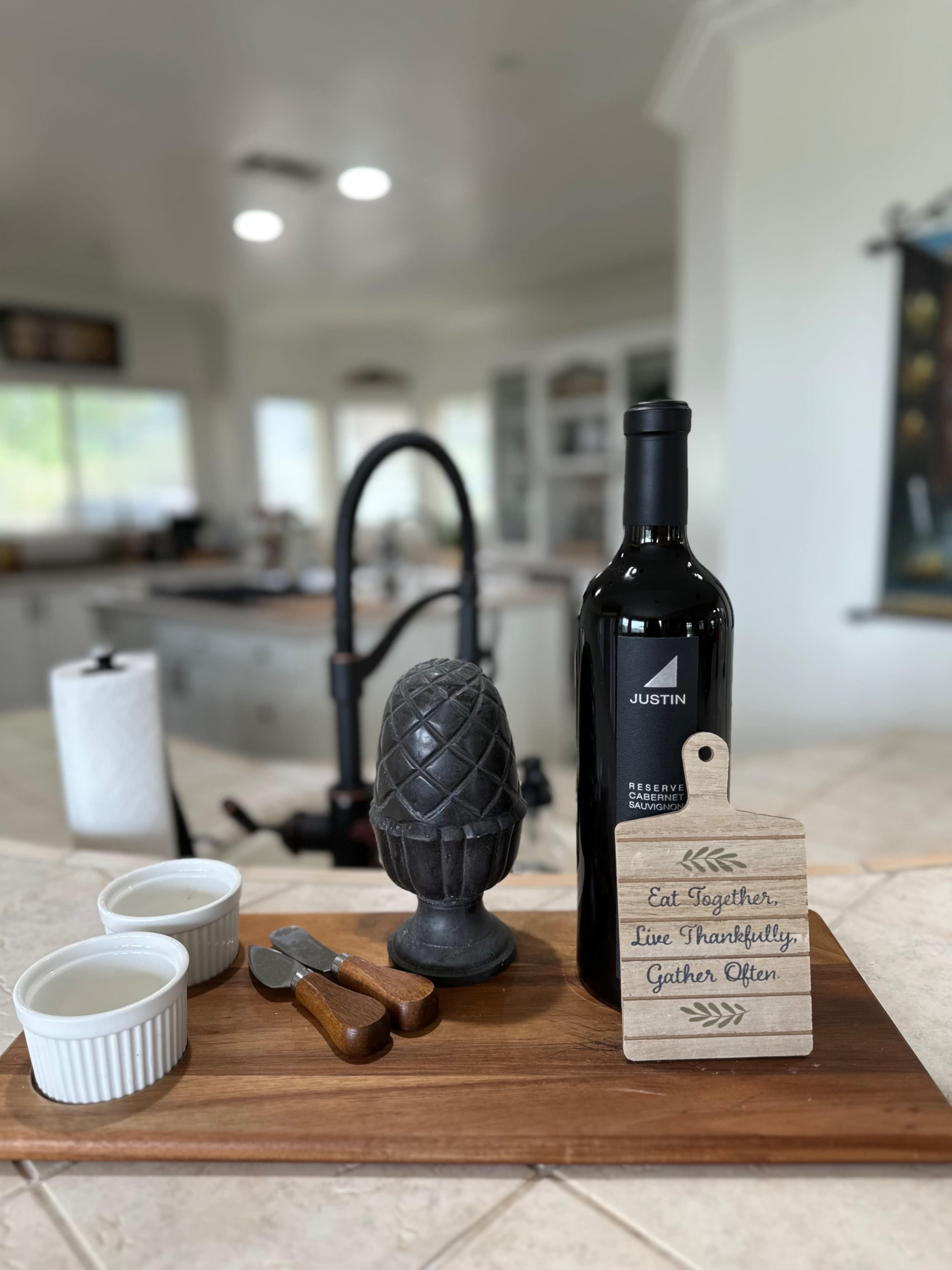 A wooden serving tray holds two white ramekins, a bottle of Justin wine, a decorative pineapple, a corkscrew, and a small sign with a motivational message, all set in a kitchen.