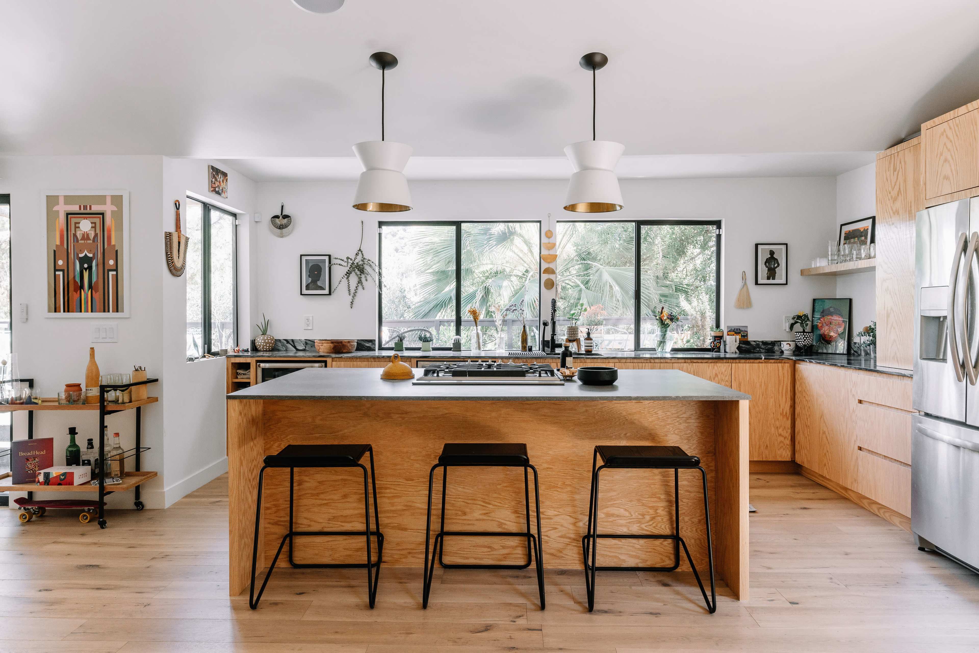 The kitchen features a large wooden island with black stools, surrounded by large windows and a mix of modern and decorative elements.
