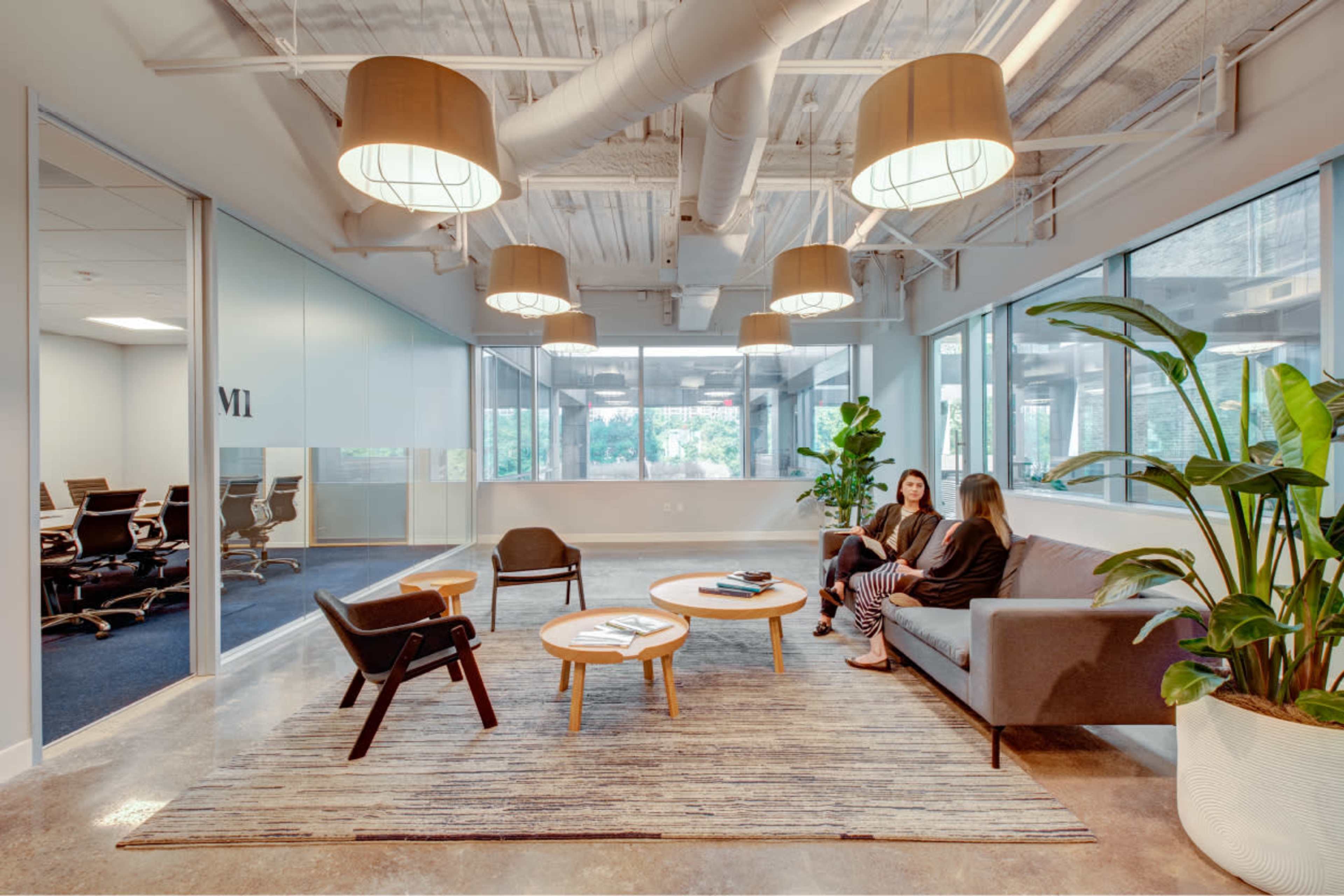 Two women sit on a couch in a modern office lobby with large windows, plants, and a waiting area featuring a coffee table and rug.