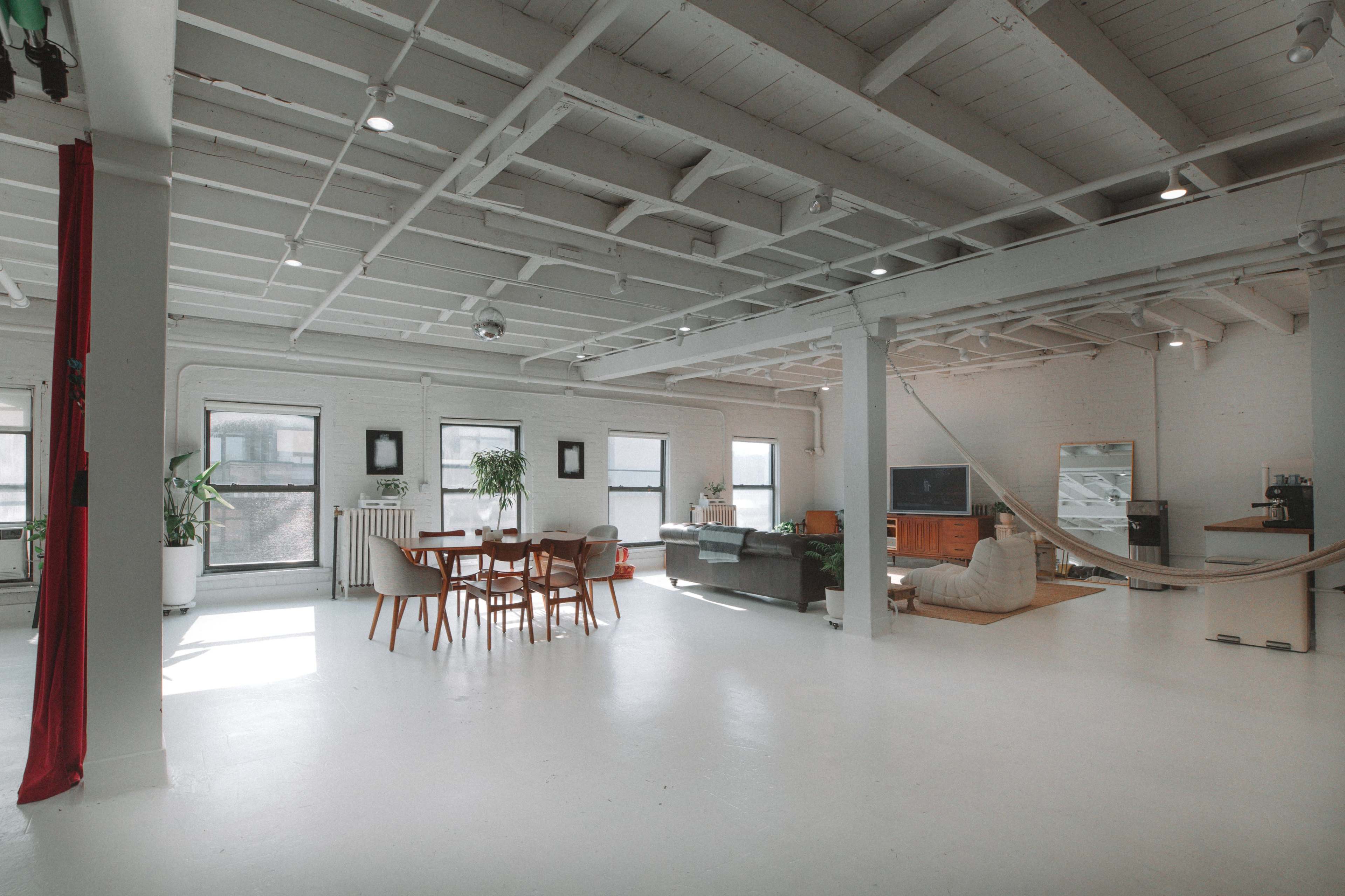 The image shows a spacious, minimalist interior with a dining area, living room, and a hammock, all featuring white walls and a bright, open layout.