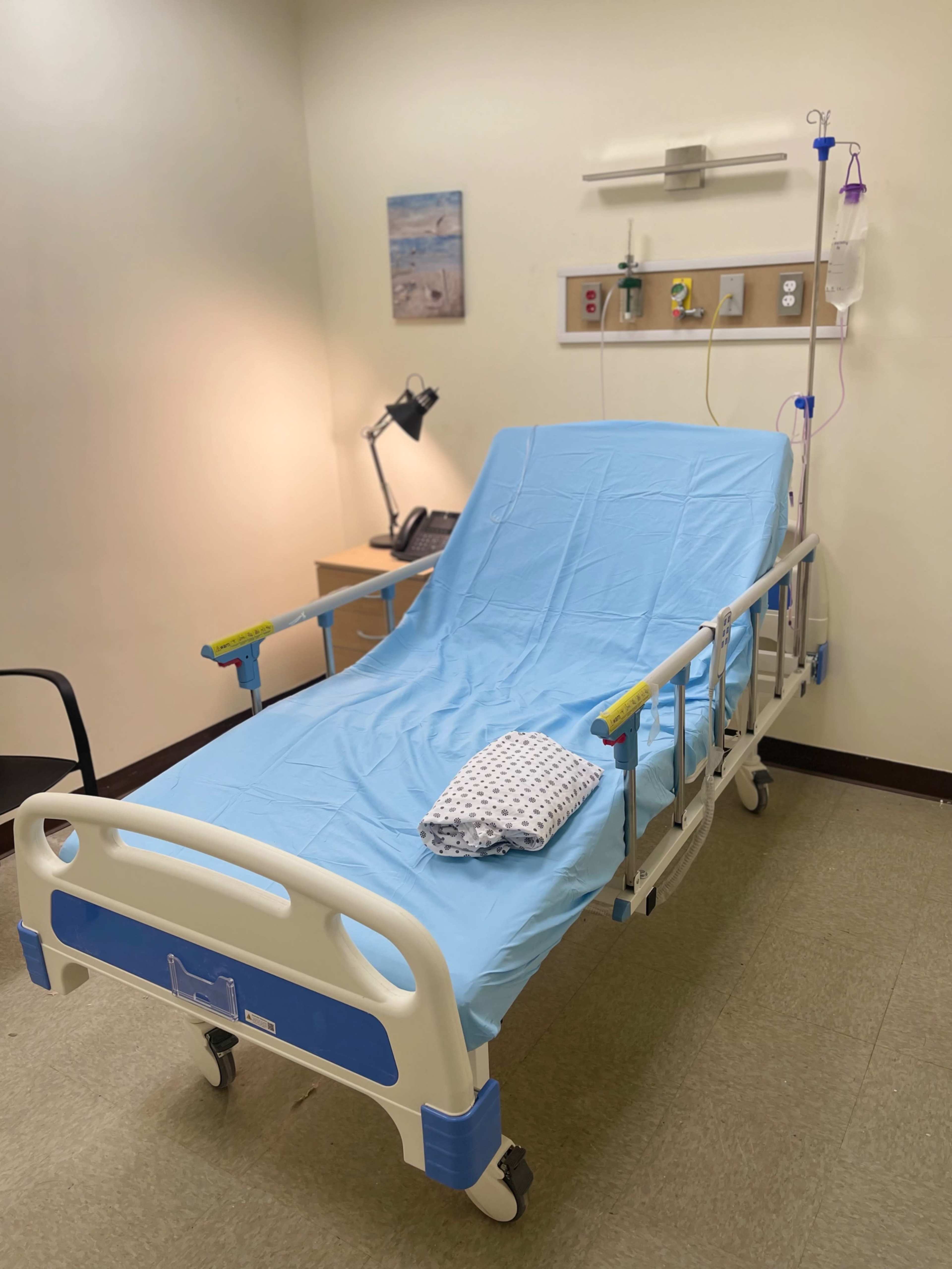 A hospital room features a medical bed with a blue sheet, a chair, and a lamp against a light-colored wall.