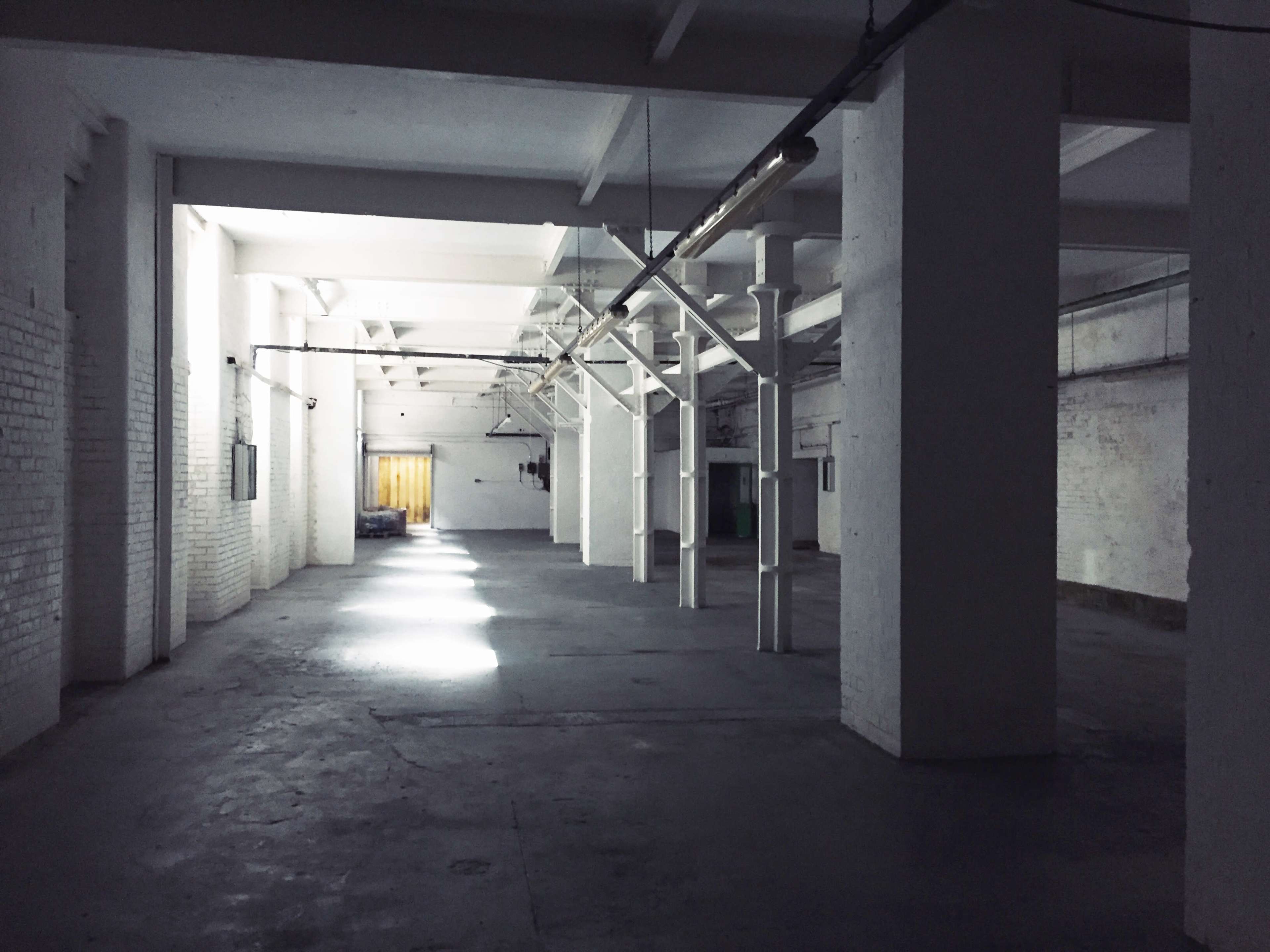 A spacious, empty industrial hall features white walls, concrete flooring, and shafts of light illuminating the interior.