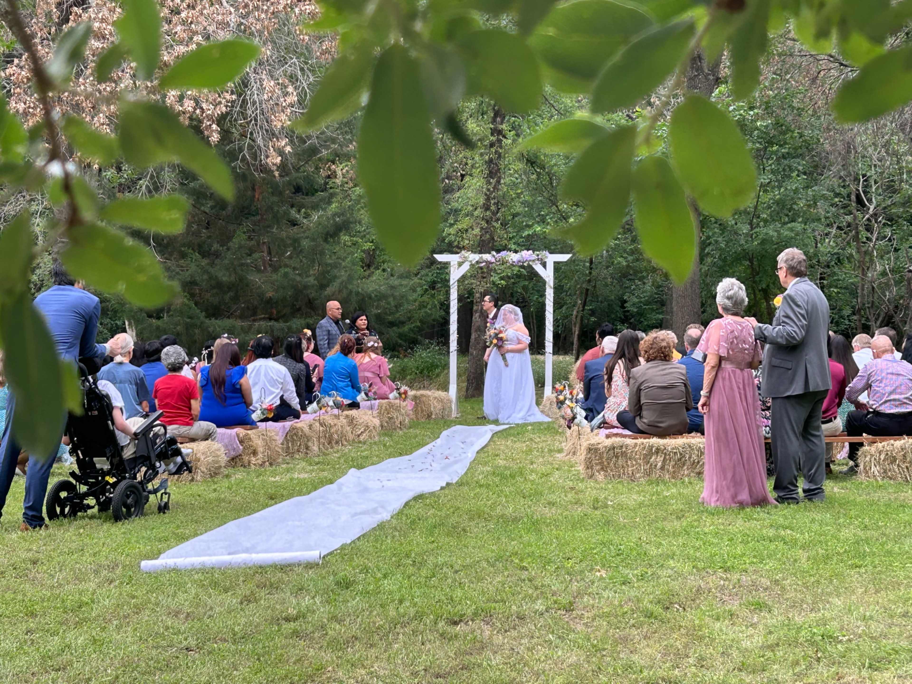 A bride walks down a grassy aisle toward a decorated archway in a garden setting, with guests seated on bales of hay.