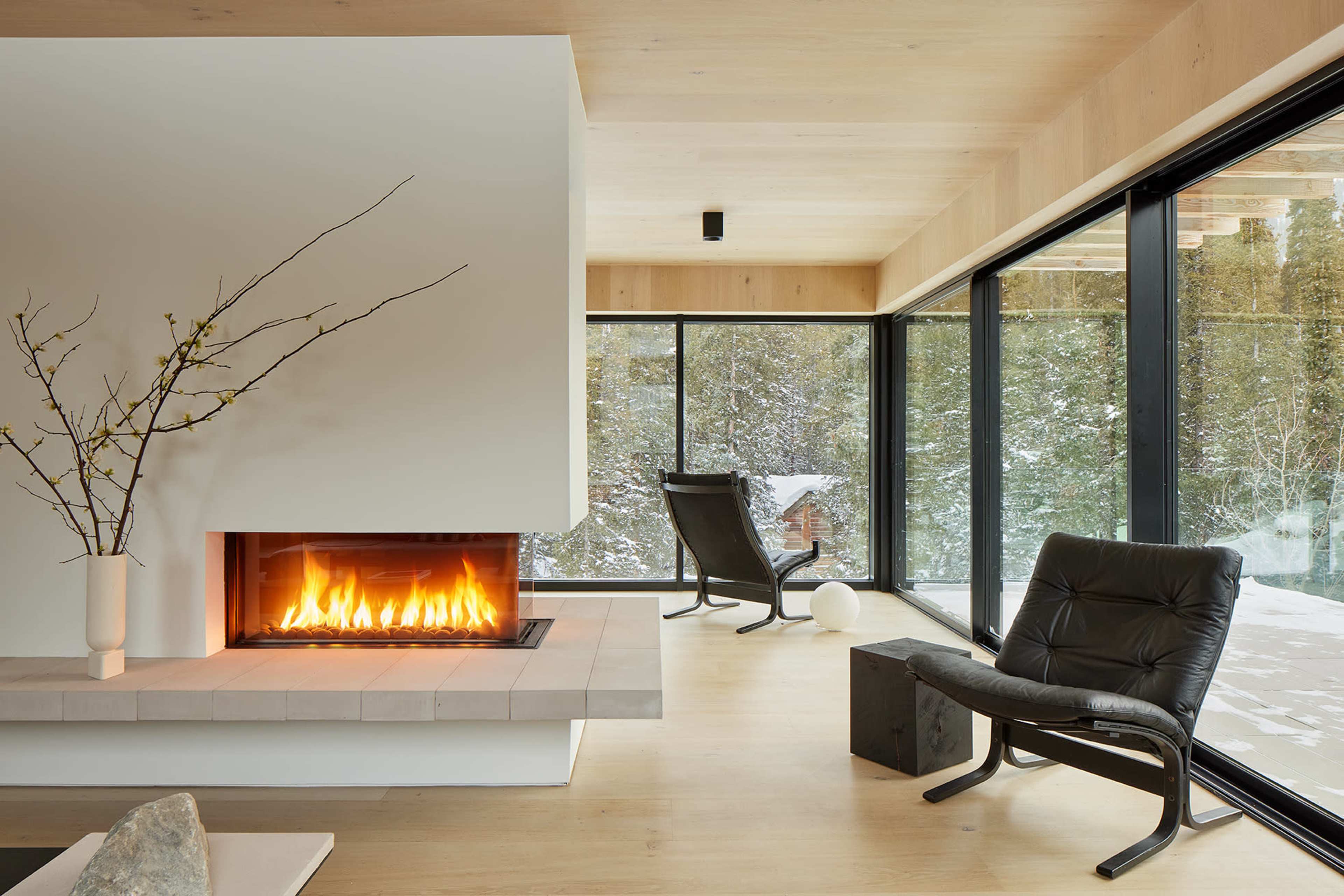A modern living room features a minimalist fireplace, large glass windows overlooking a snowy landscape, and contemporary seating arrangements.