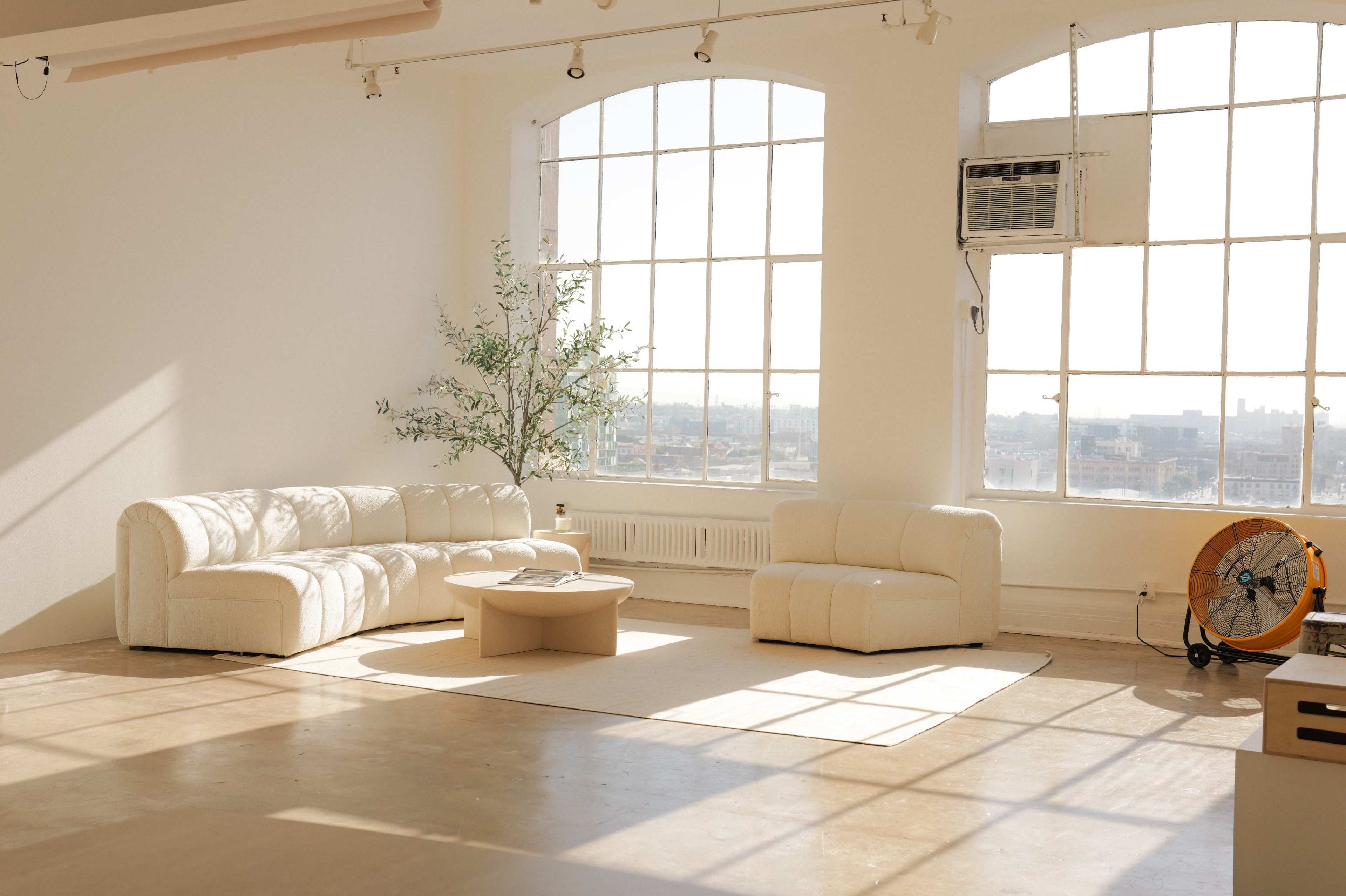The image shows a bright, minimalist living space featuring a white couch, an armchair, and a circular table, with large windows allowing natural light to enter.