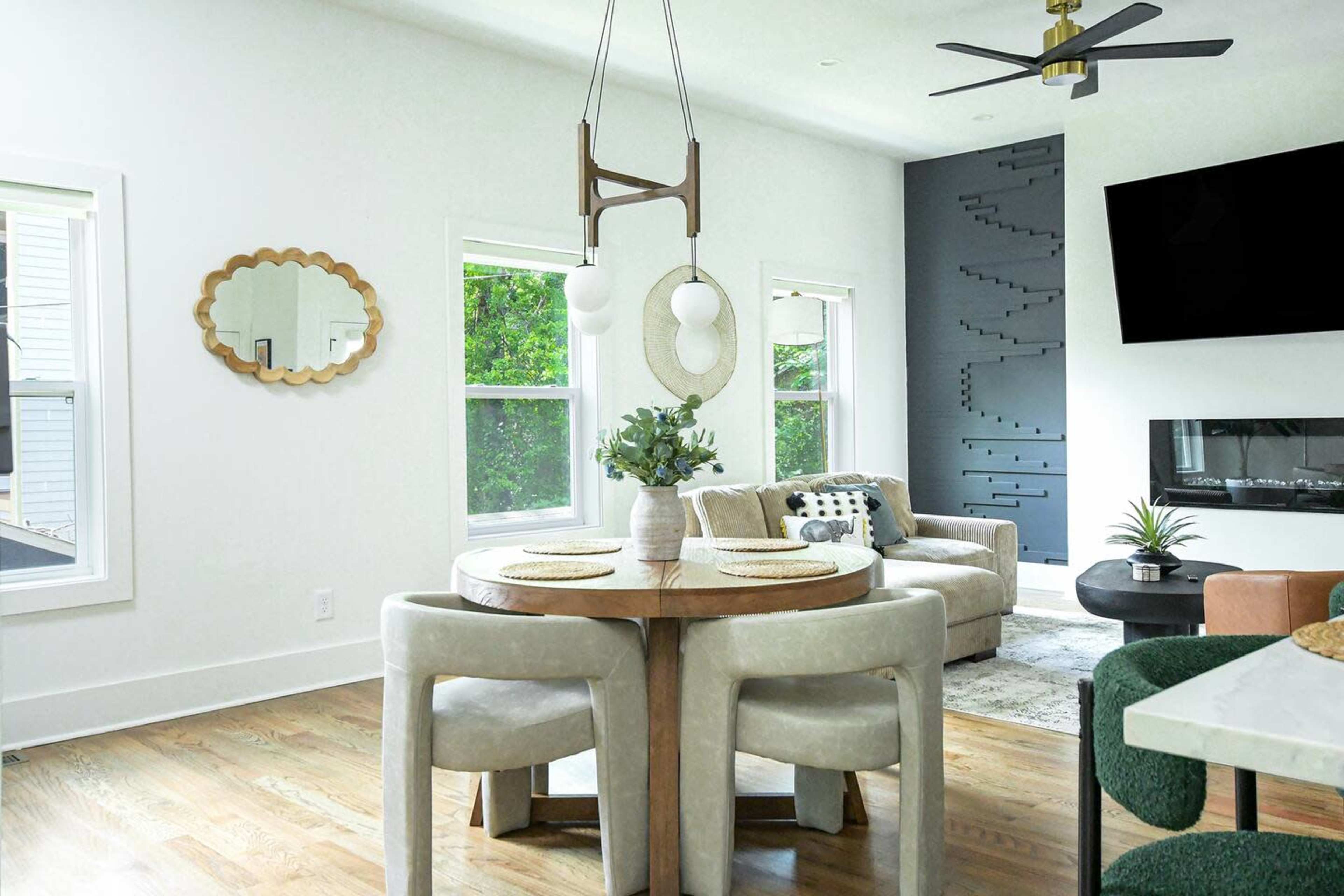 The image shows a modern dining area with a round wooden table, upholstered chairs, and a wall-mounted TV next to a cozy seating arrangement.