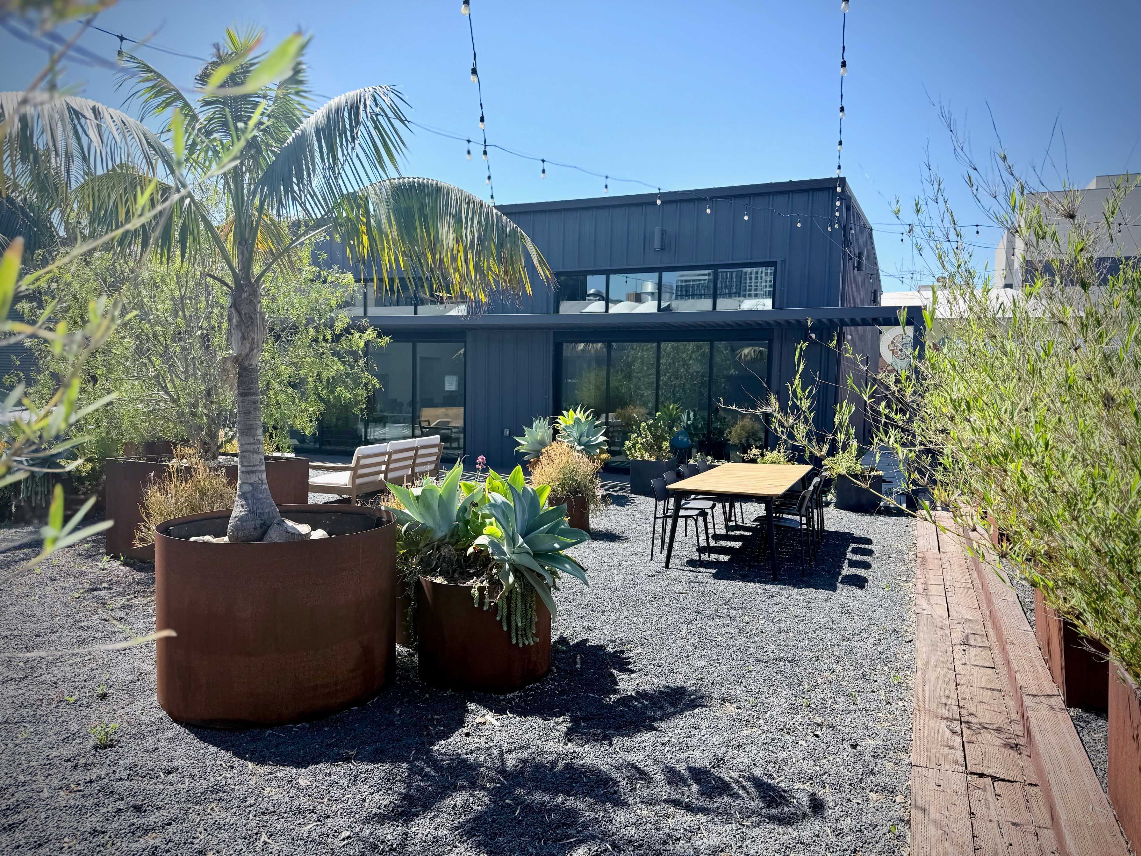 The image shows a modern outdoor space featuring several large planters, a long dining table, and a building in the background under a clear blue sky.