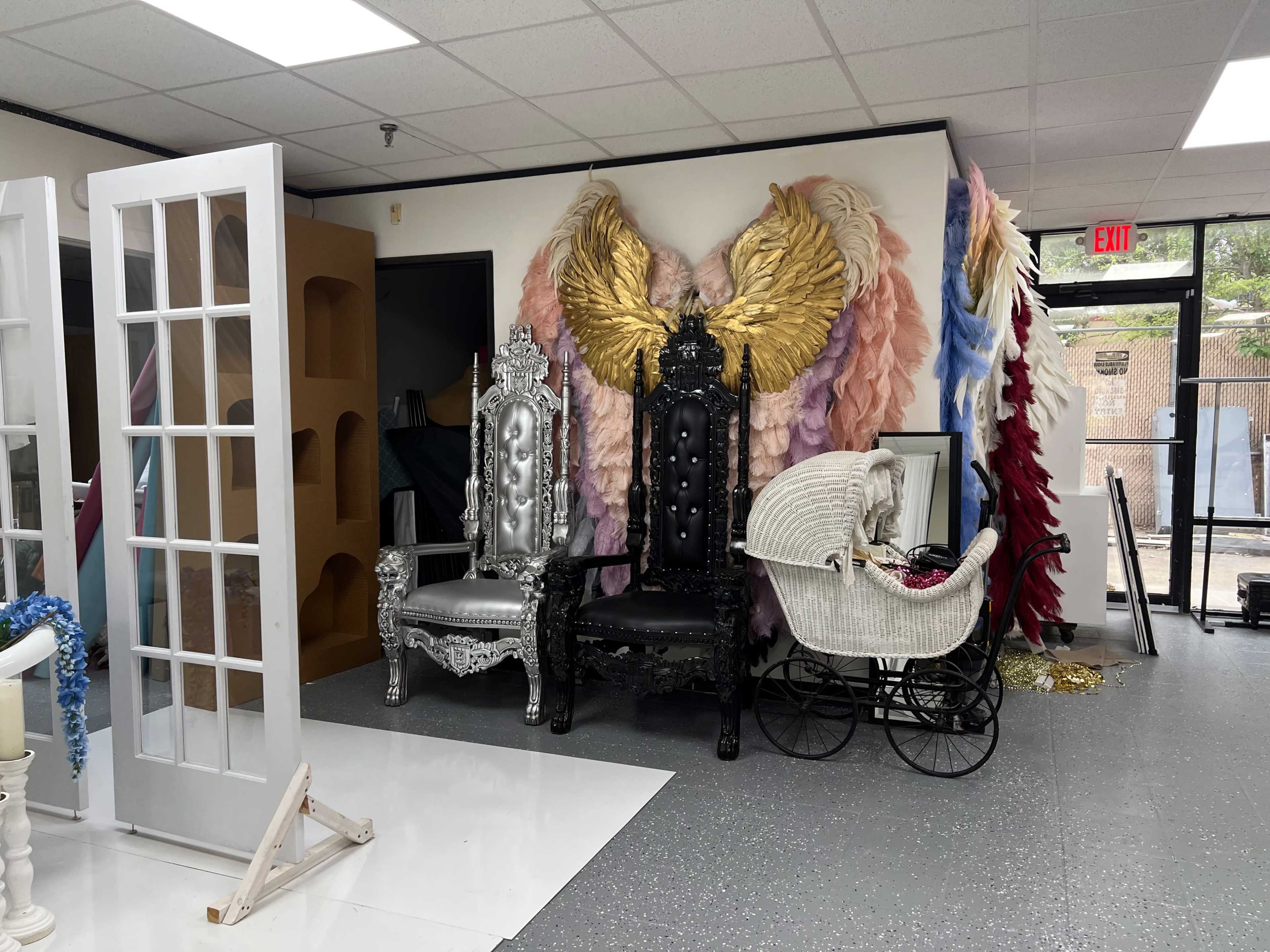 The image shows a decorated room featuring two large ornate chairs, one silver and one black, positioned against a wall adorned with large, colorful angel wings made of fabric.